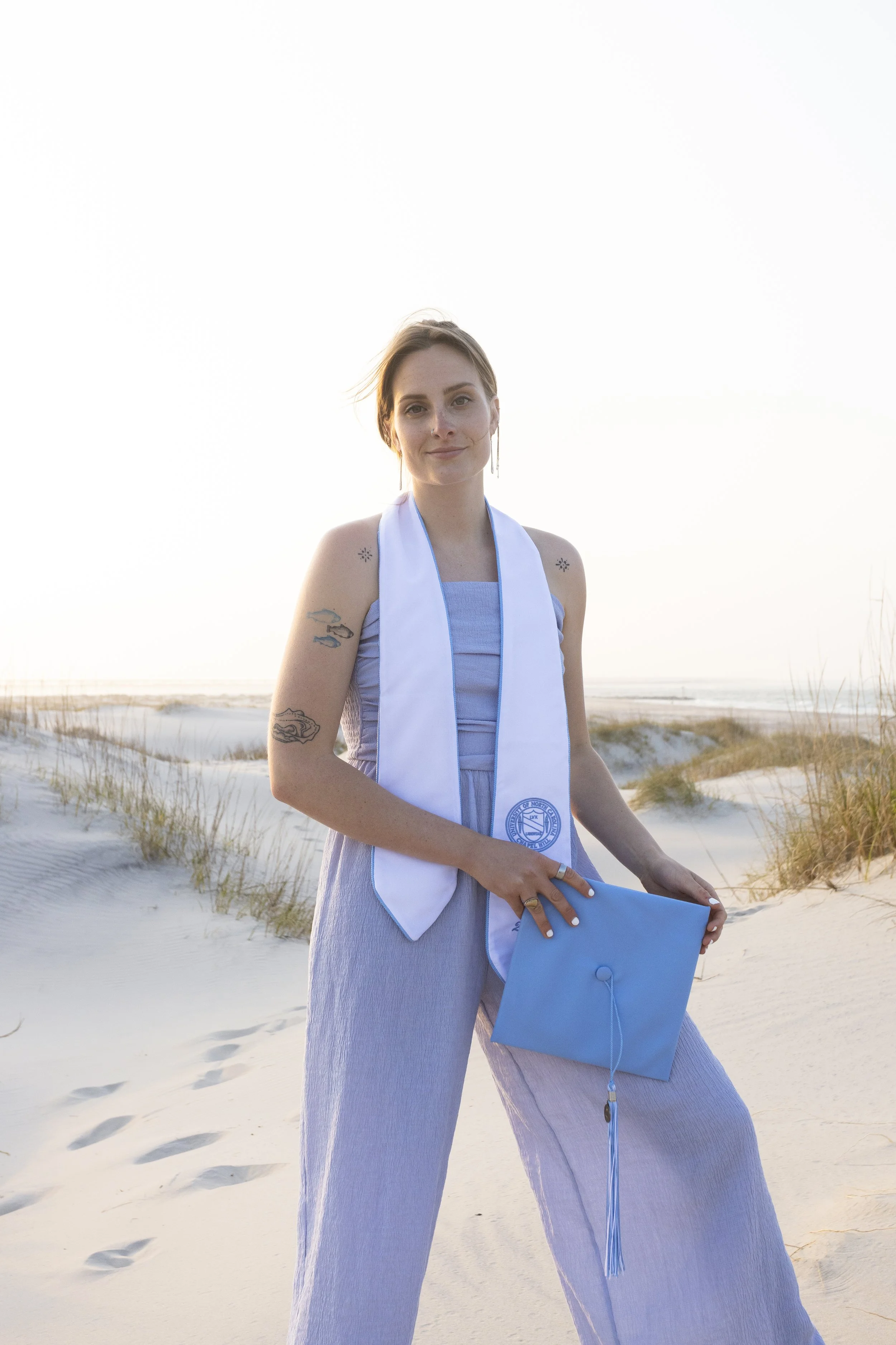 A young woman standing on a sandy beach during sunset, wearing a light gray dress, a white stole with a blue emblem, and holding a blue diploma cover.