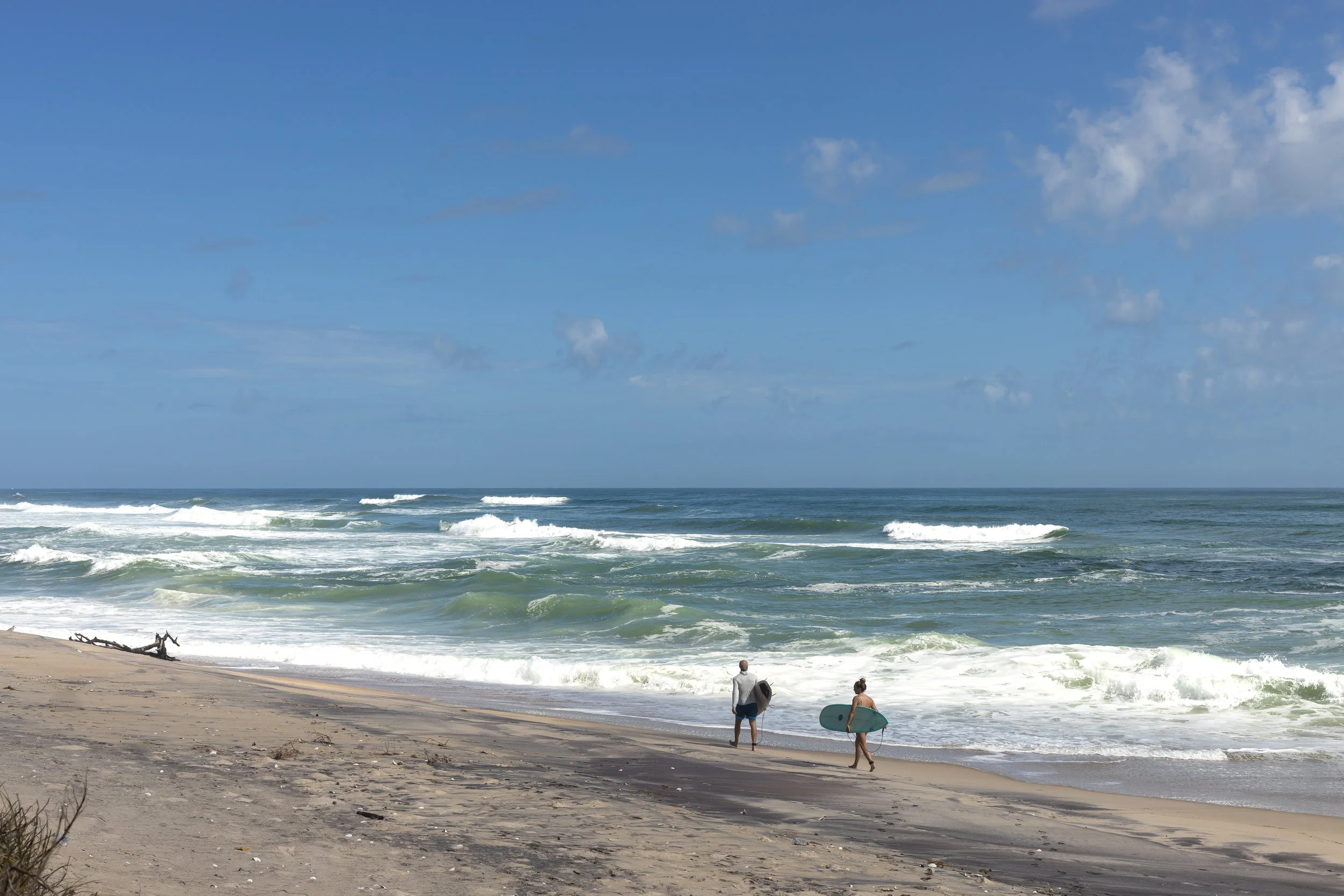 Two surfers walking on the sandy beach towards the ocean, carrying surfboards, under a partly cloudy sky.