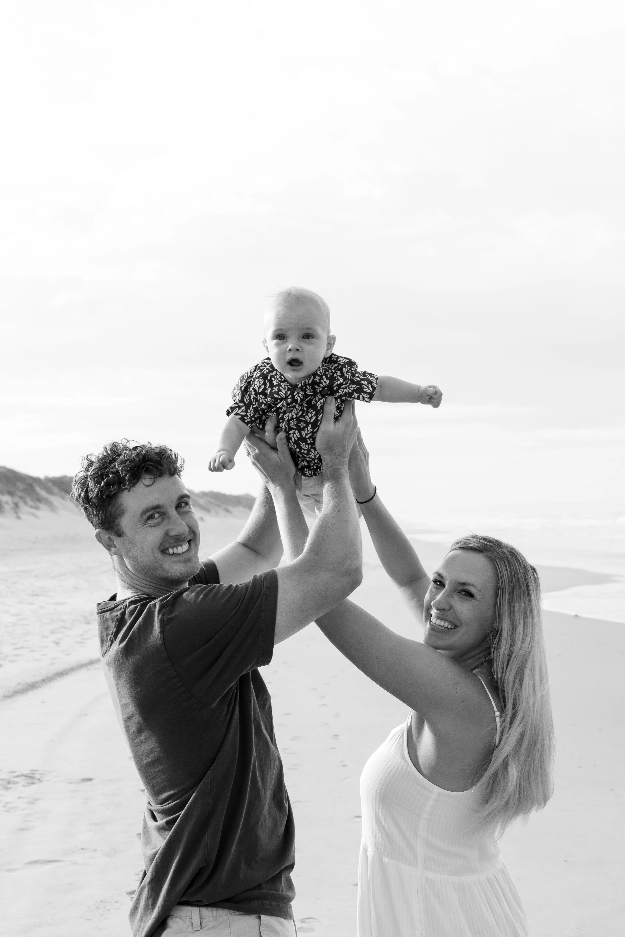 A happy family of three at the beach, with the father and mother lifting their baby into the air, all smiling and enjoying the moment.