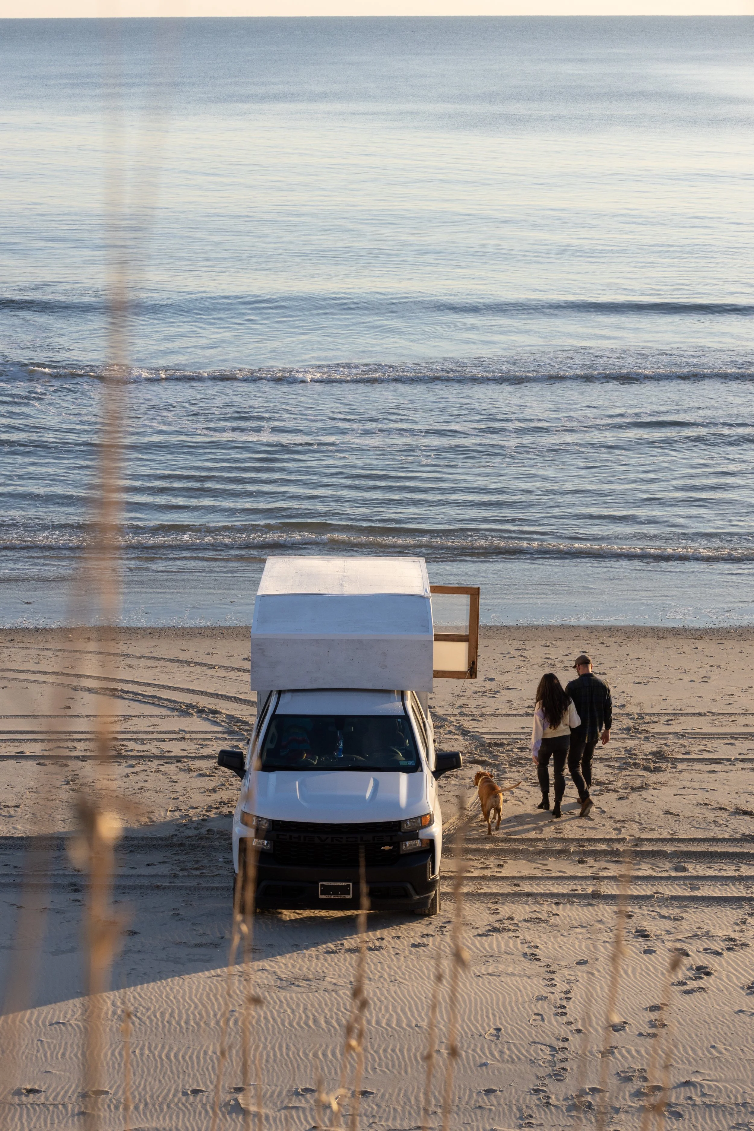 A white camper van parked on a sandy beach facing the ocean, with two people and a dog walking along the shoreline at sunset.