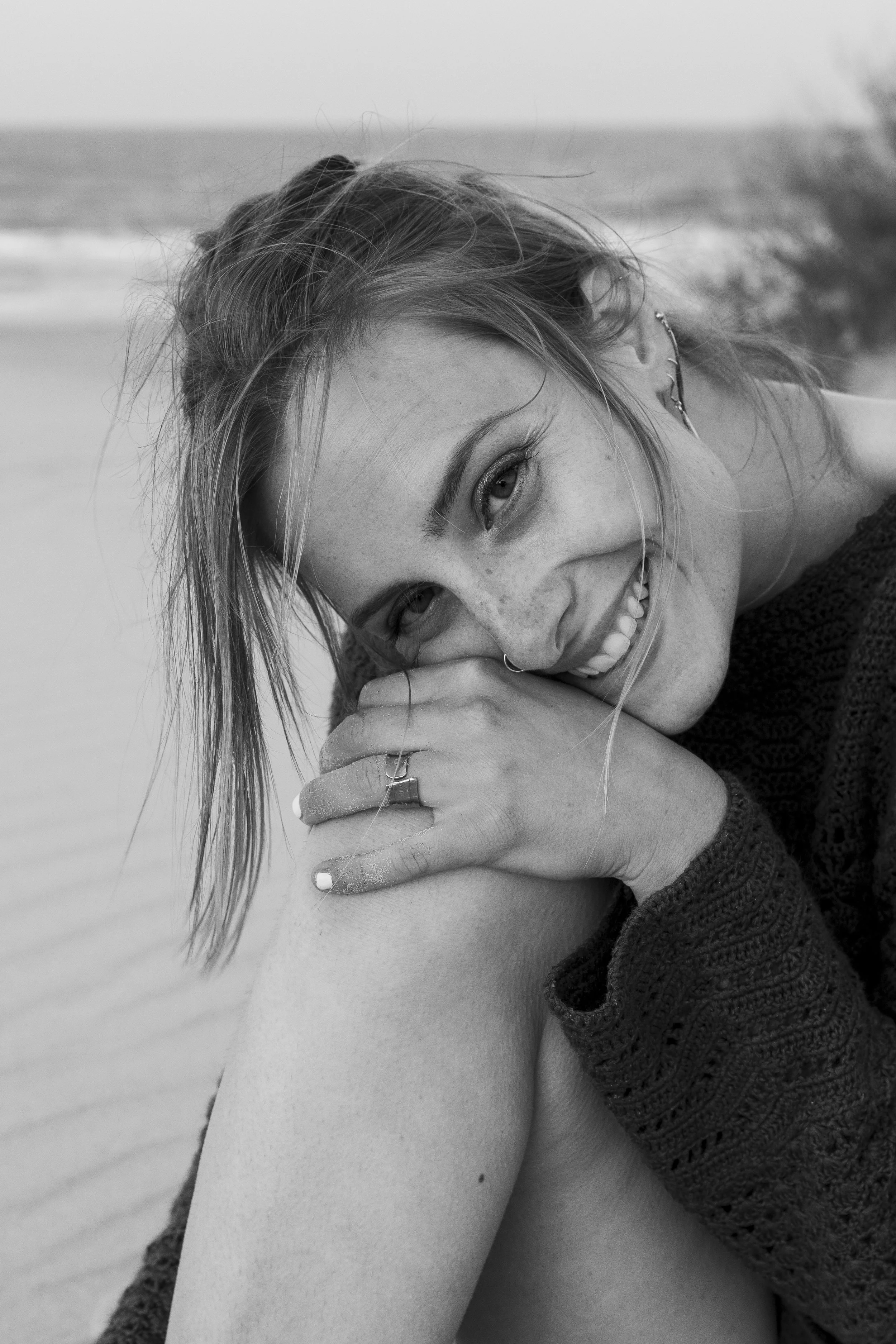 Black and white photograph of a smiling woman with freckles, lying on her side on a beach, resting her head on her knee, with ocean waves in the background.