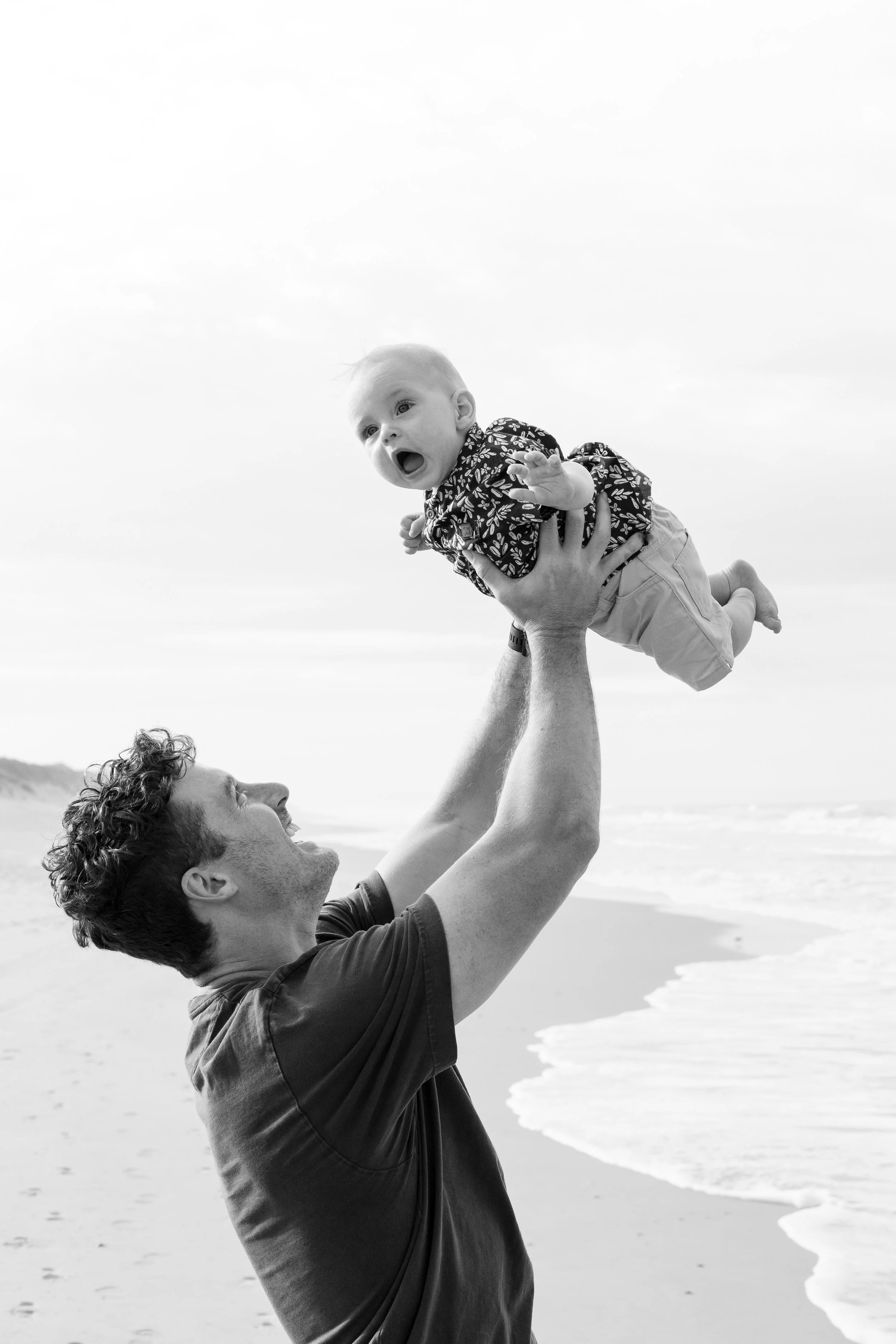 A man at the beach lifting a baby into the air, both smiling and enjoying the moment.
