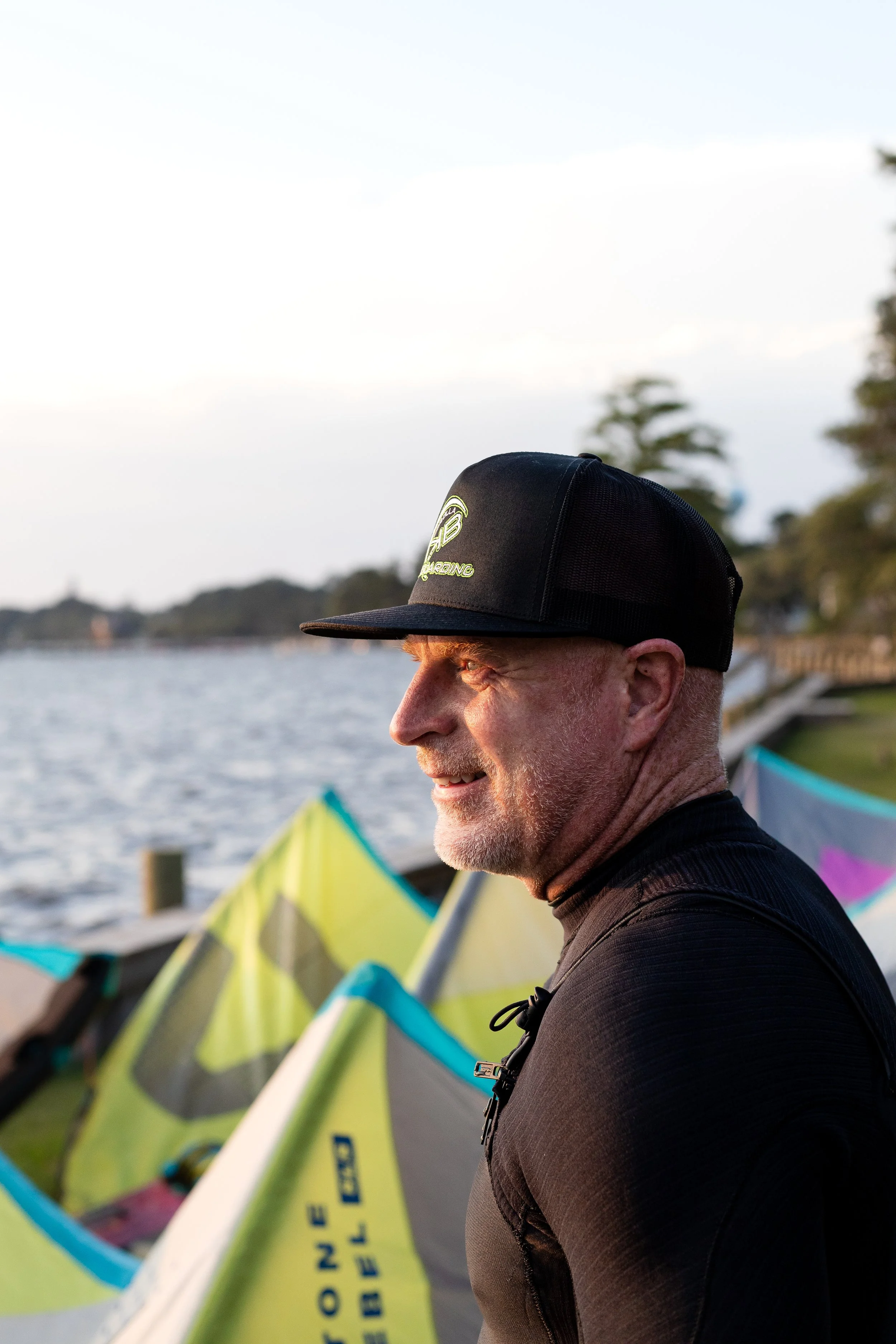 A close-up of a smiling man wearing a black cap and black athletic shirt standing outdoors near water with colorful growlers or kites in the background, during sunset.