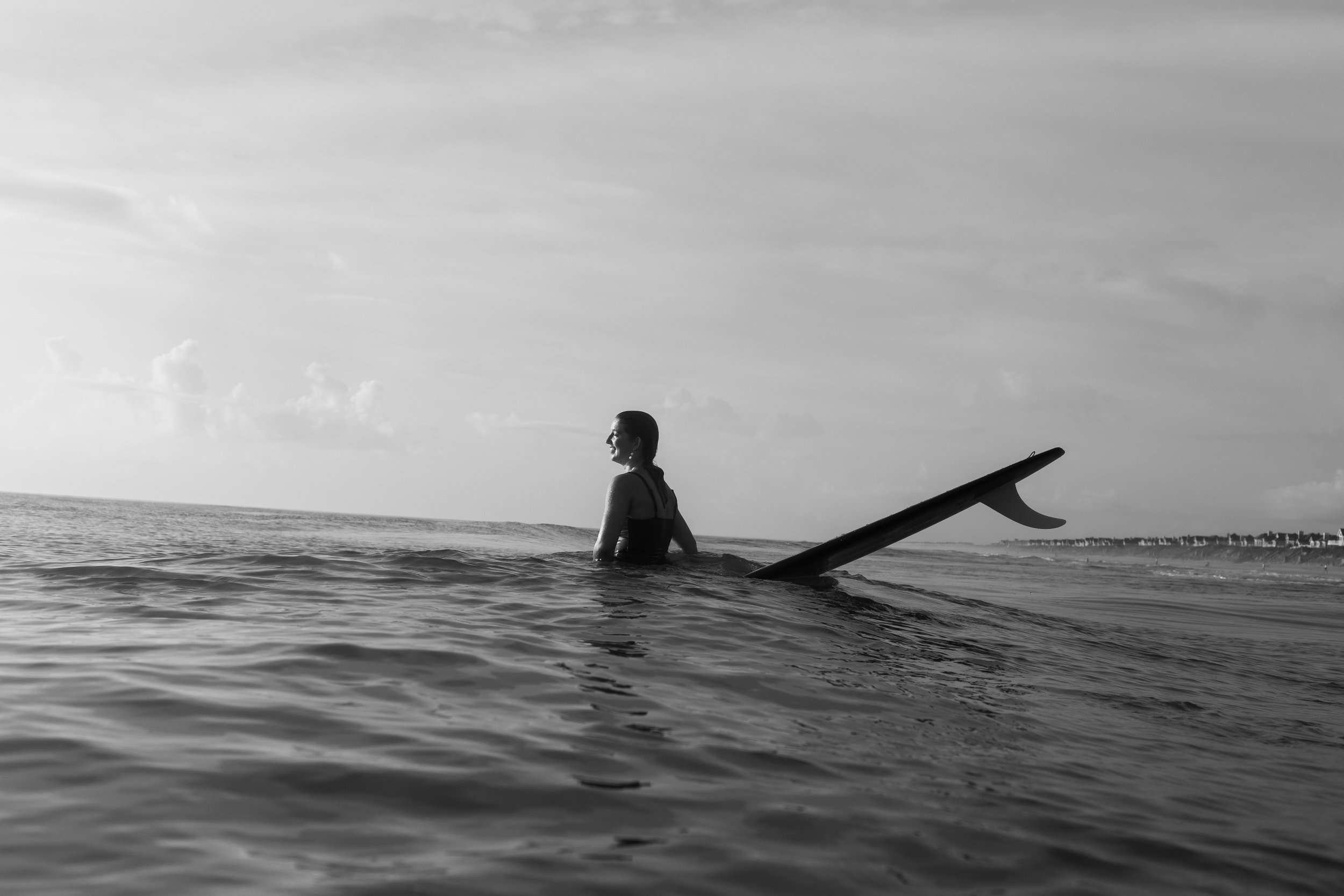 A woman is standing in the ocean with a surfboard partially submerged in the water, facing to the right, with a distant shoreline and a cloudy sky in the background.