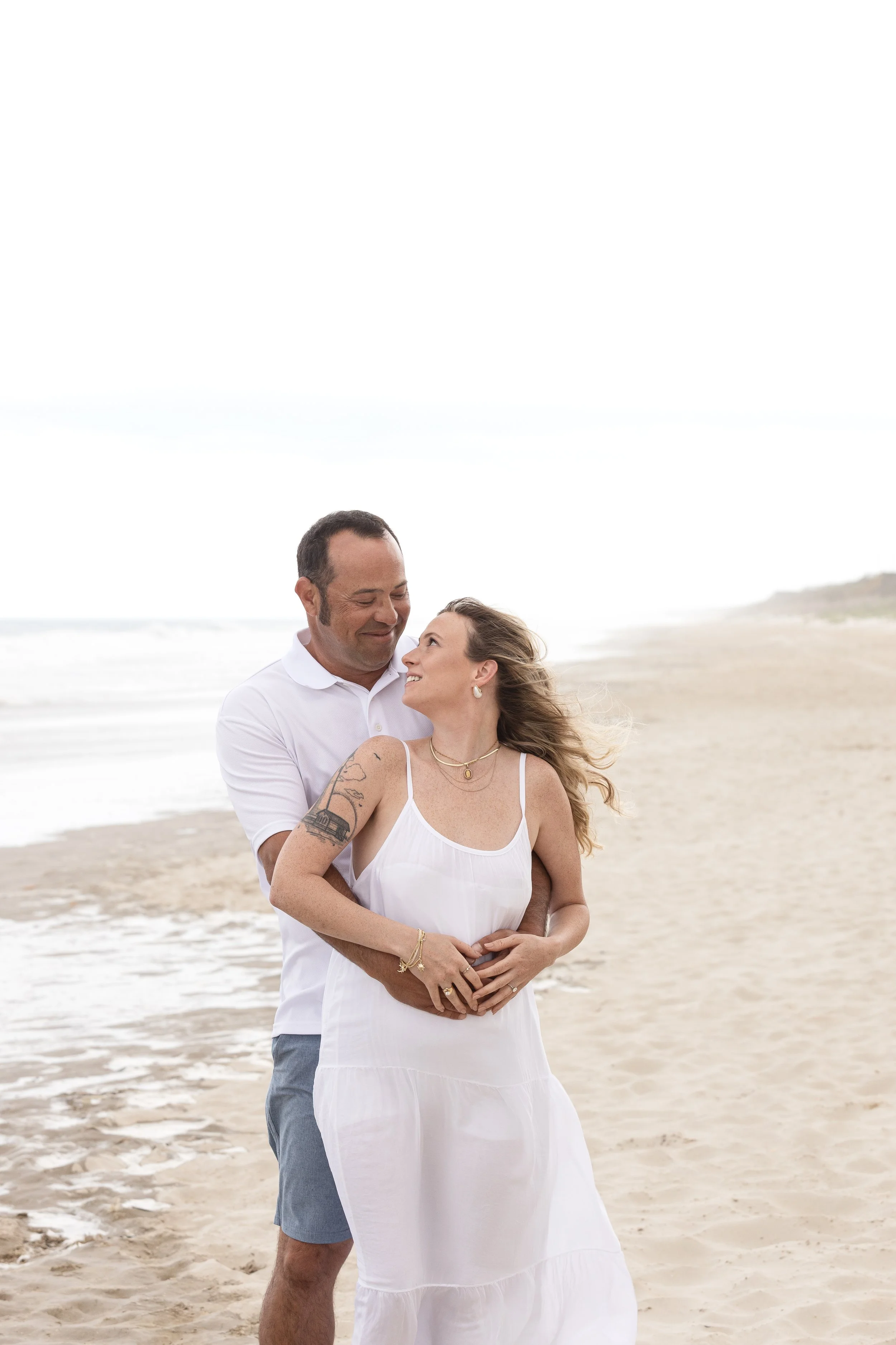 A couple standing on the beach embracing and smiling at each other. The woman has long hair, tattoos on her arm, and is wearing a white dress. The man is wearing a white shirt and blue shorts. The background features the sandy beach and ocean waves.