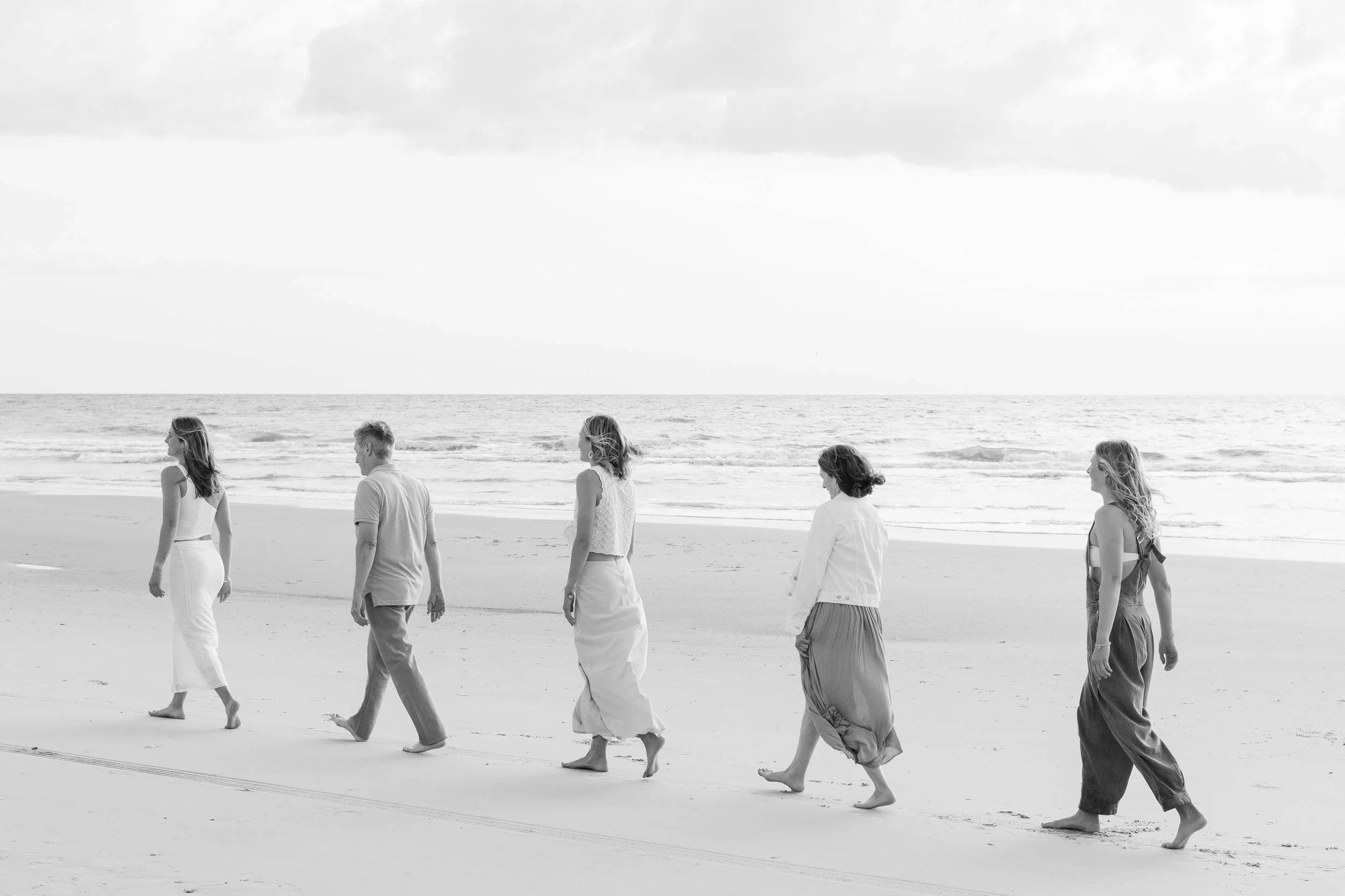 Six women and one man walking on the beach near the ocean, dressed in casual summer clothing.