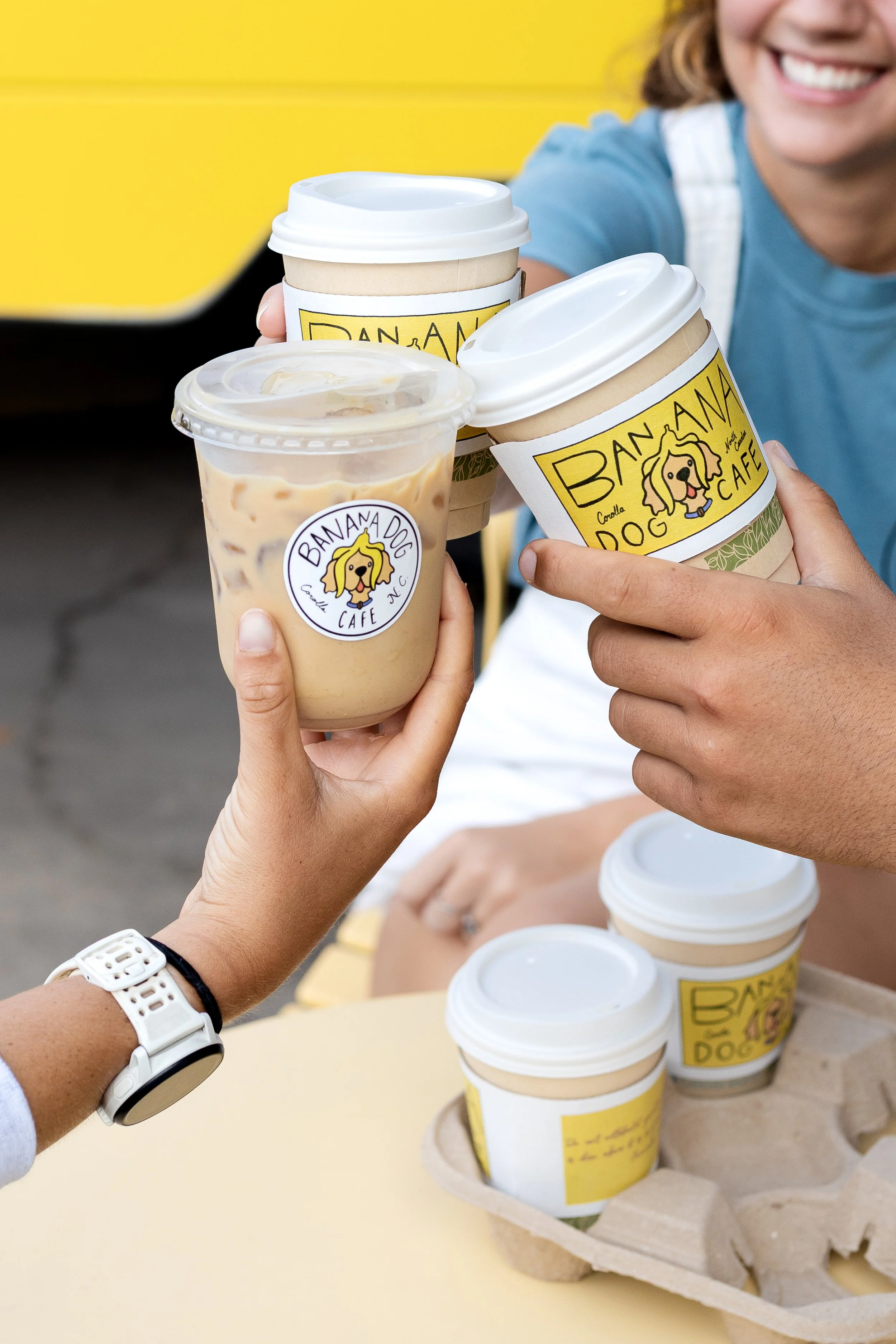 People toasting with coffee and iced drinks in a cafe with a bright yellow background.