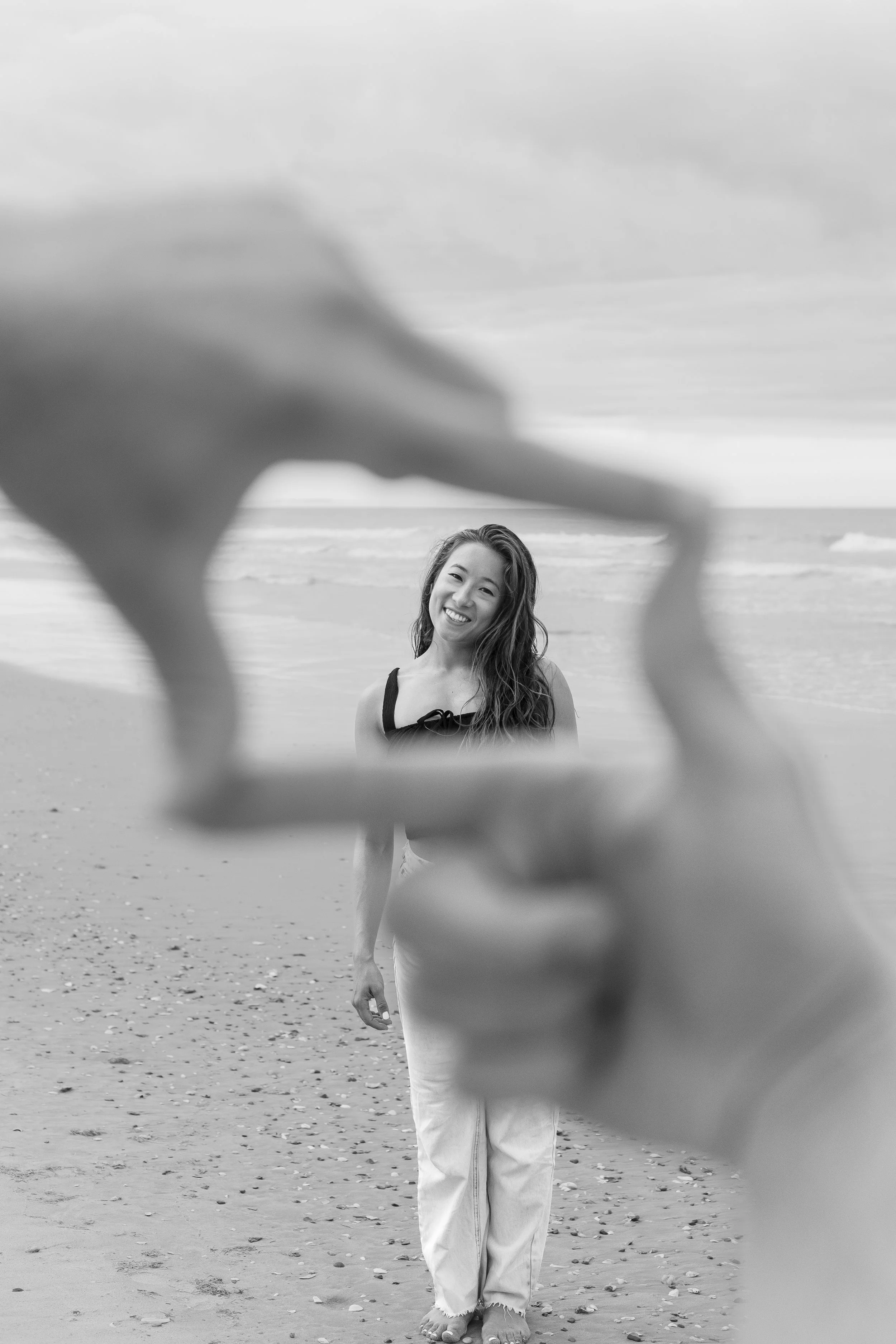 A woman standing on a beach, smiling and looking at the camera, with ocean waves and sky in the background. The photo is in black and white and is framed through a person's hand making a square.