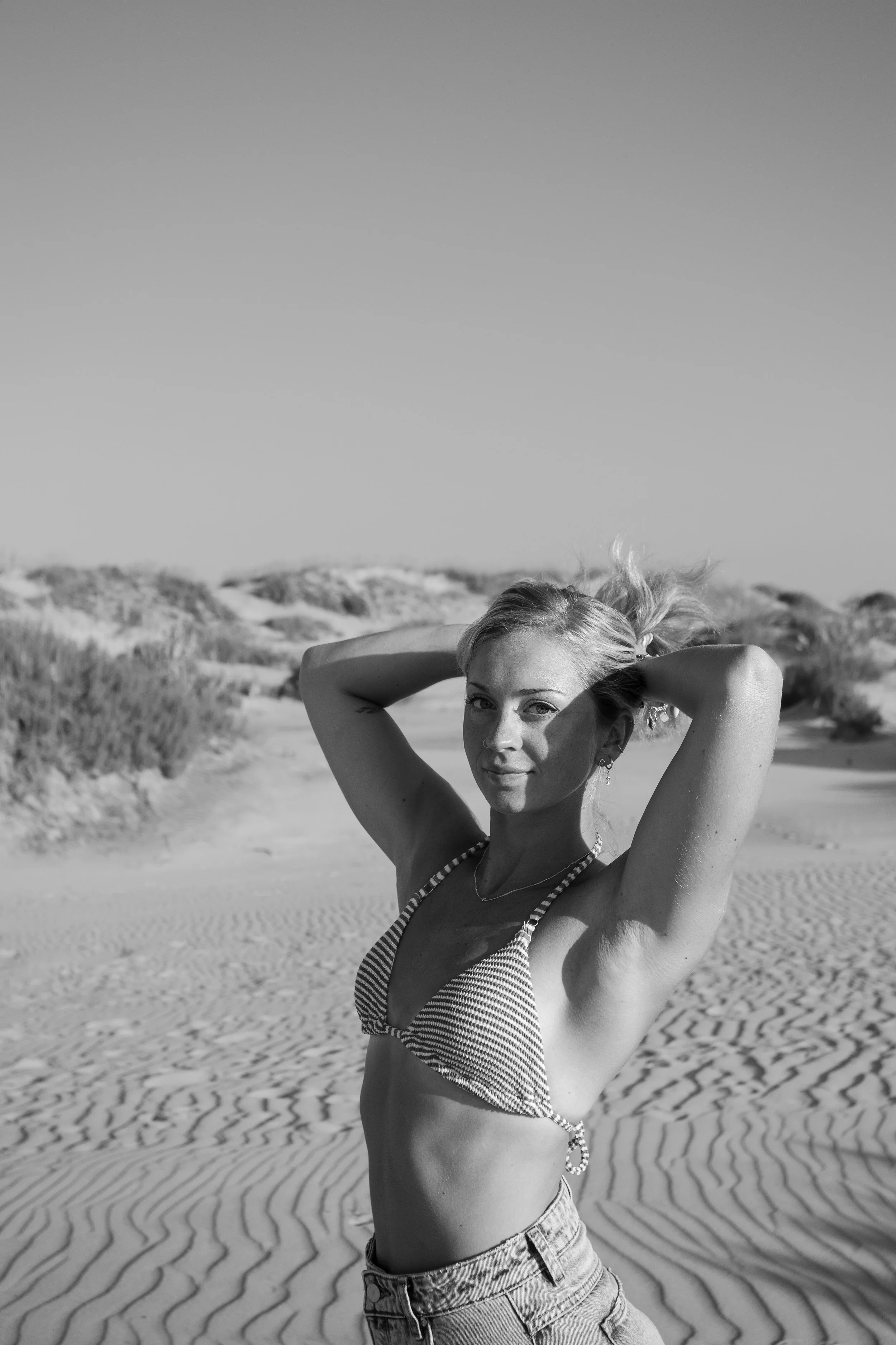 Black and white photo of a young woman at the beach with sand dunes and sparse vegetation in the background. She is wearing a striped bikini top and high-waisted shorts, with her arms raised behind her head, smiling slightly at the camera.