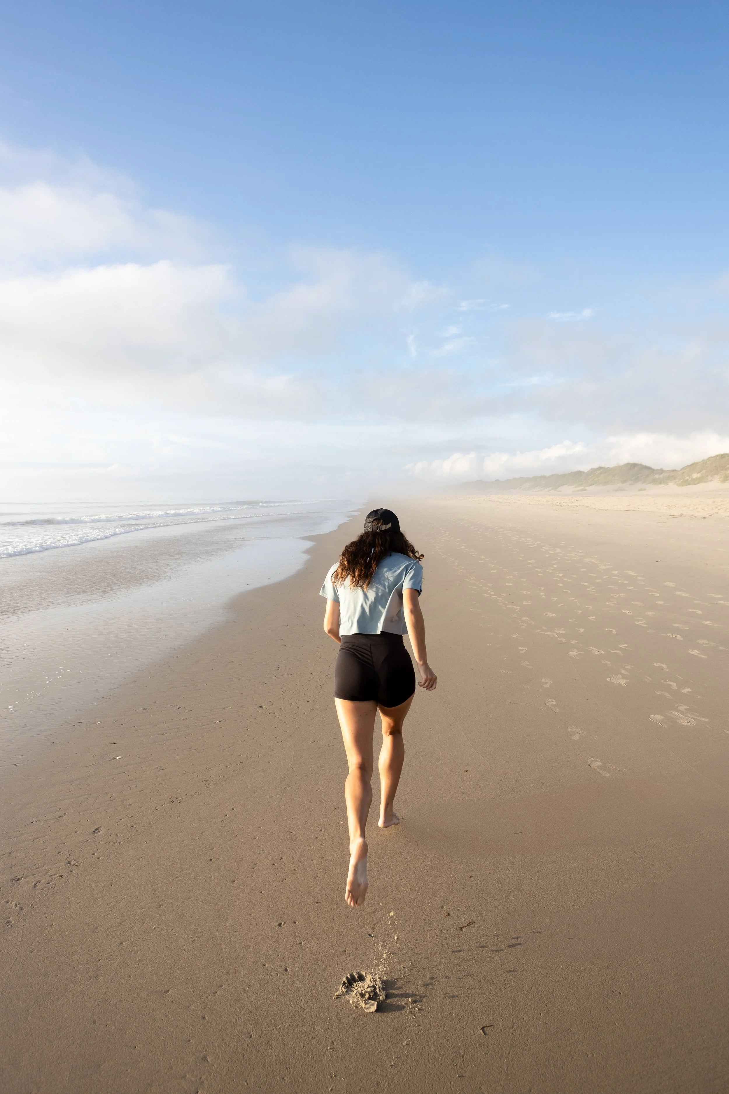 A woman running barefoot on a sandy beach, leaving footprints, with a blue sky and ocean waves in the background.