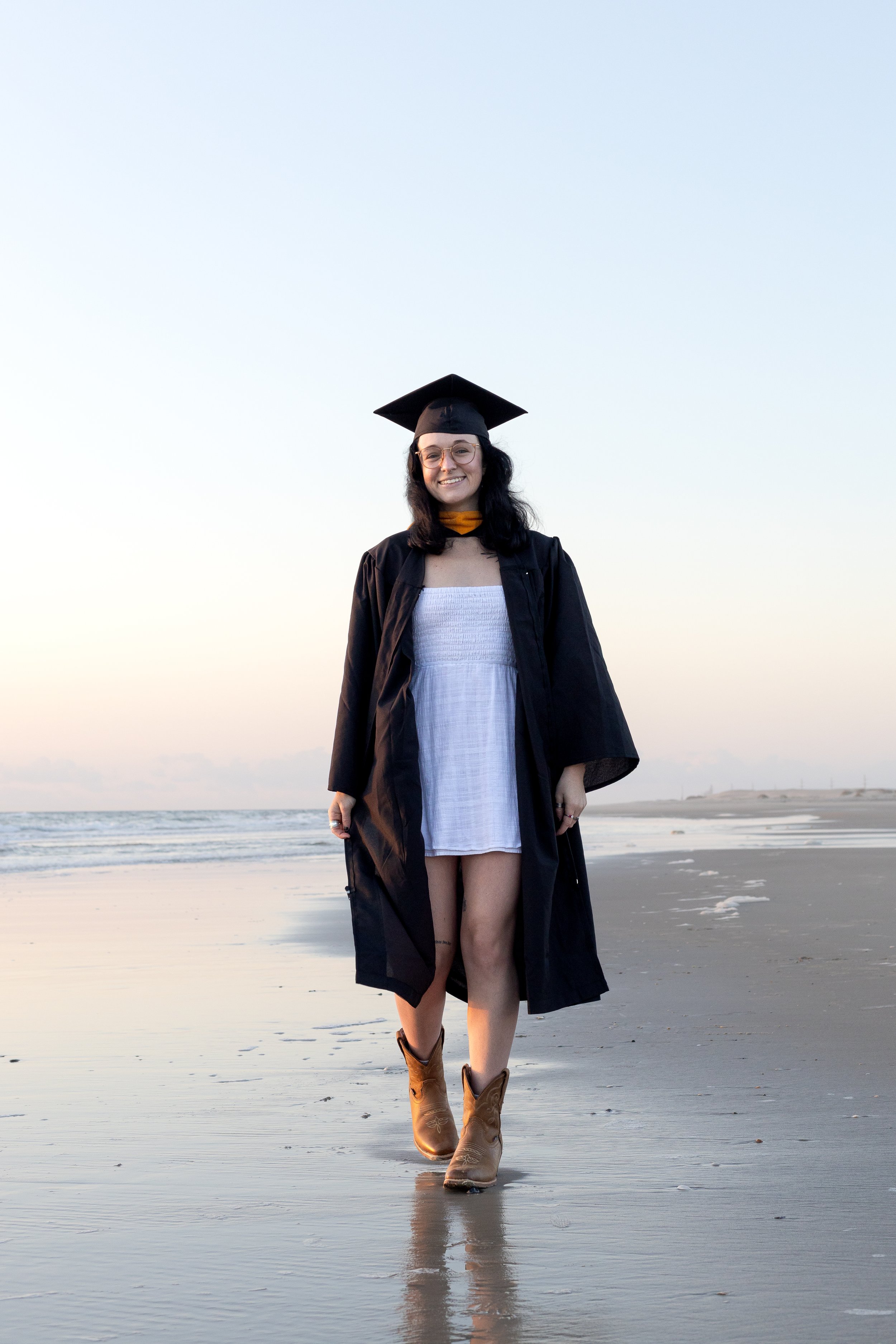 A woman in a graduation cap and gown walking on a beach during sunset.