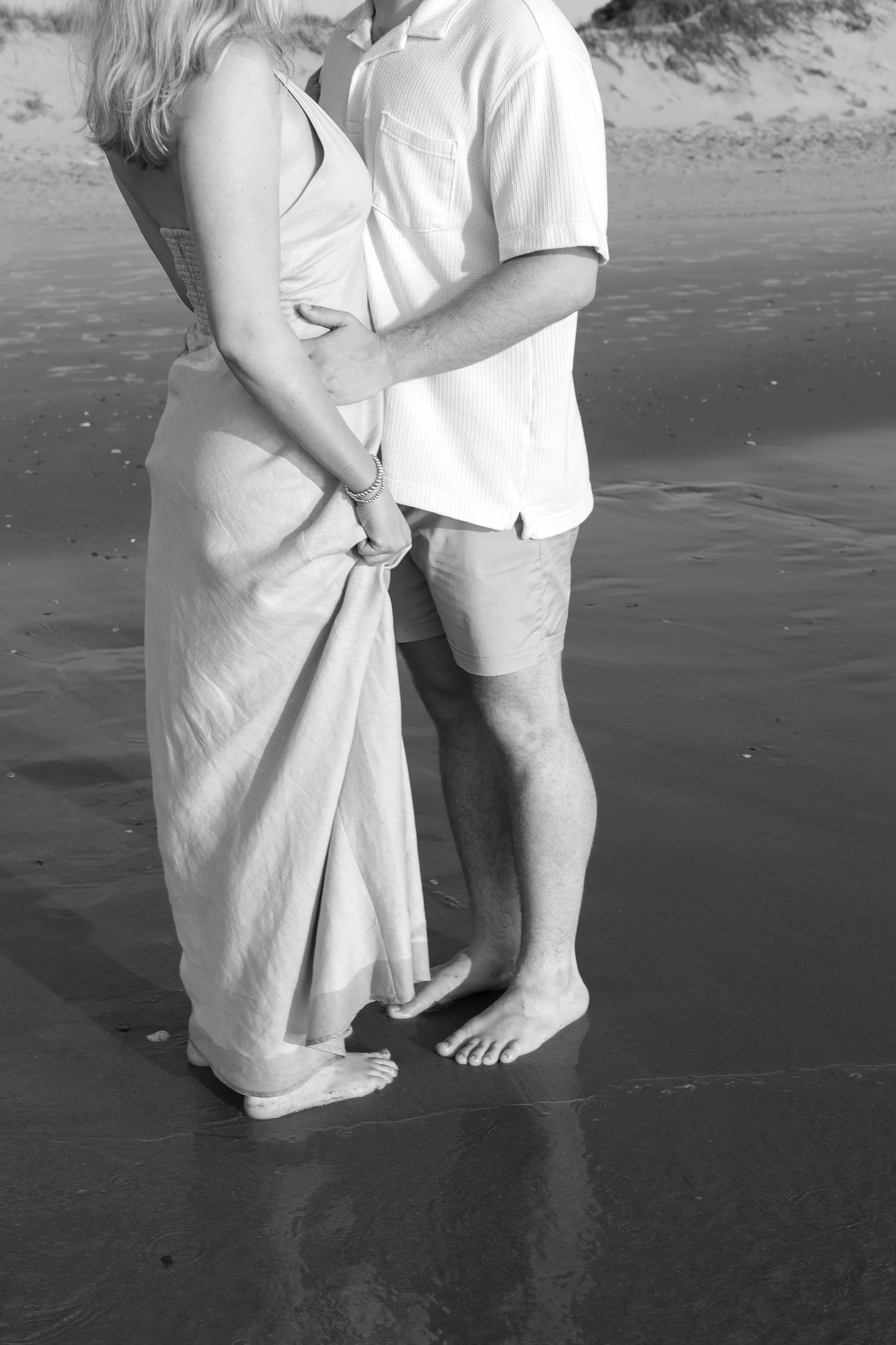 A couple standing close together on a beach, holding hands and embracing barefoot in the sand, with ocean waves in the background.
