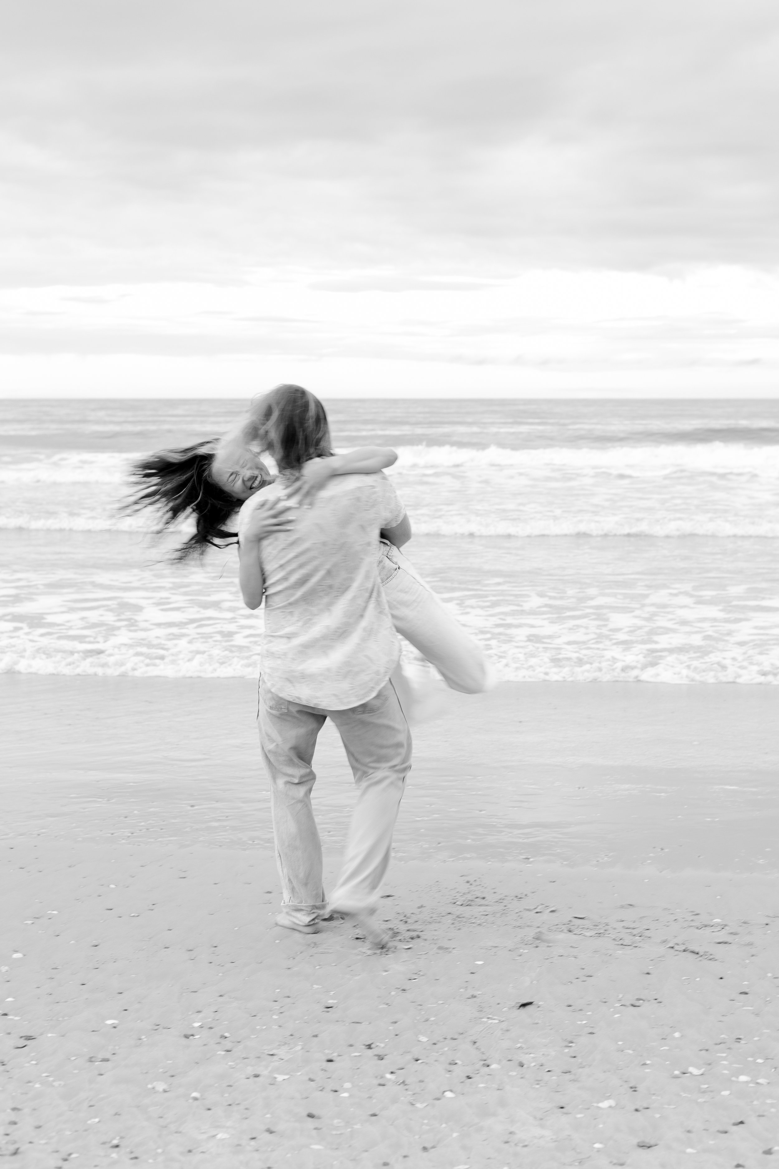 Two people, one adult and one child, sharing a joyful hug on the beach with waves and cloudy sky in the background.