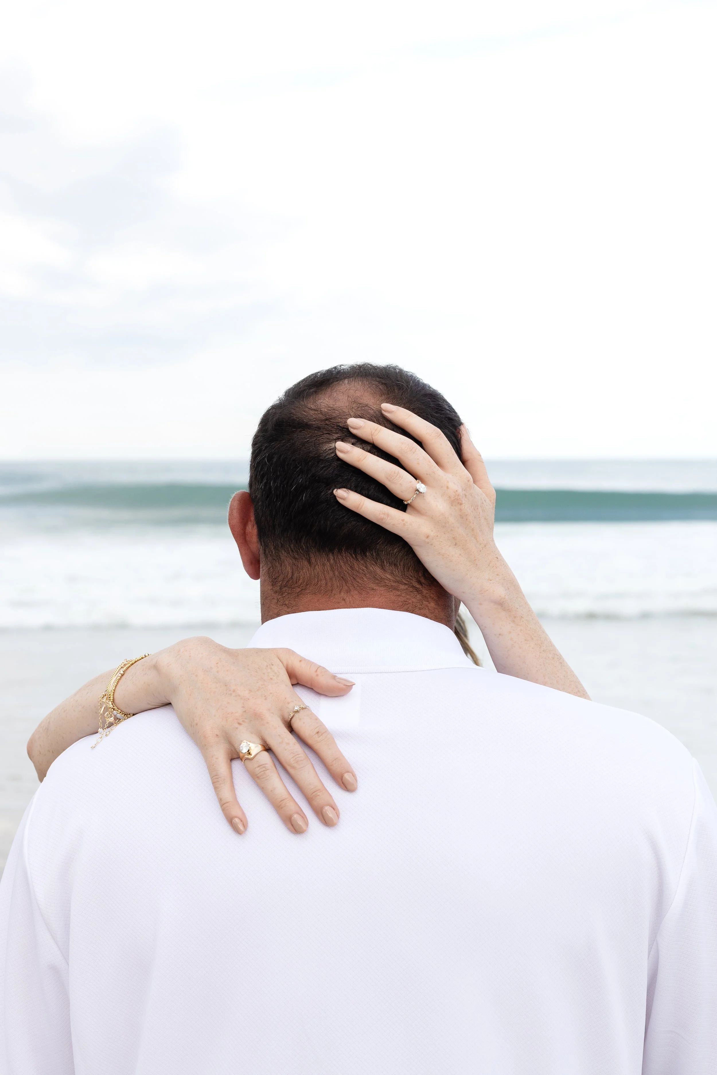 Couple embracing at the beach, with the woman's hand on the man's head and her other arm around his neck, showing wedding rings and jewelry.