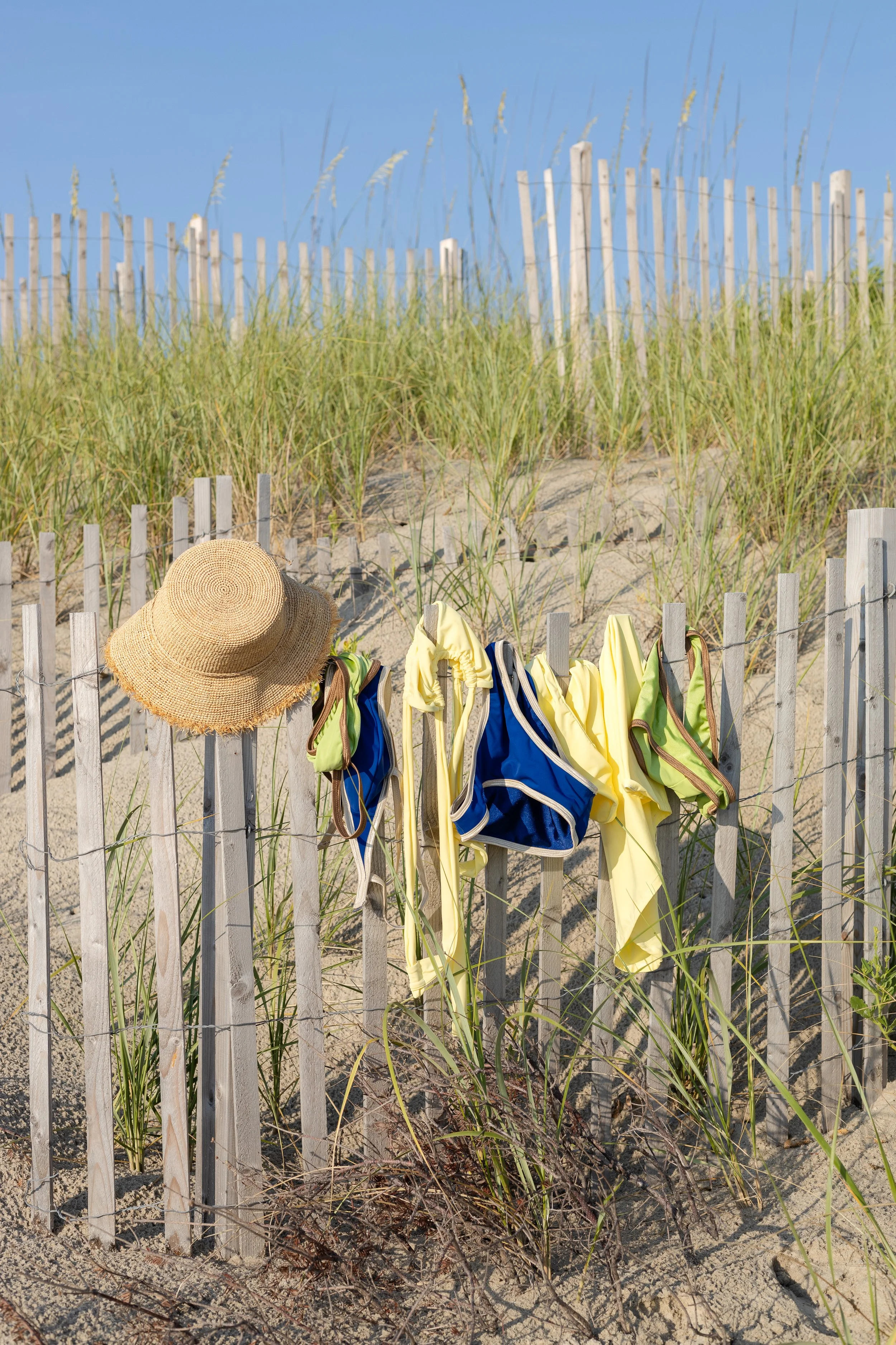 Beach fence with a sun hat and colorful swimsuits hanging on it, sand with grass, and a blue sky in the background.
