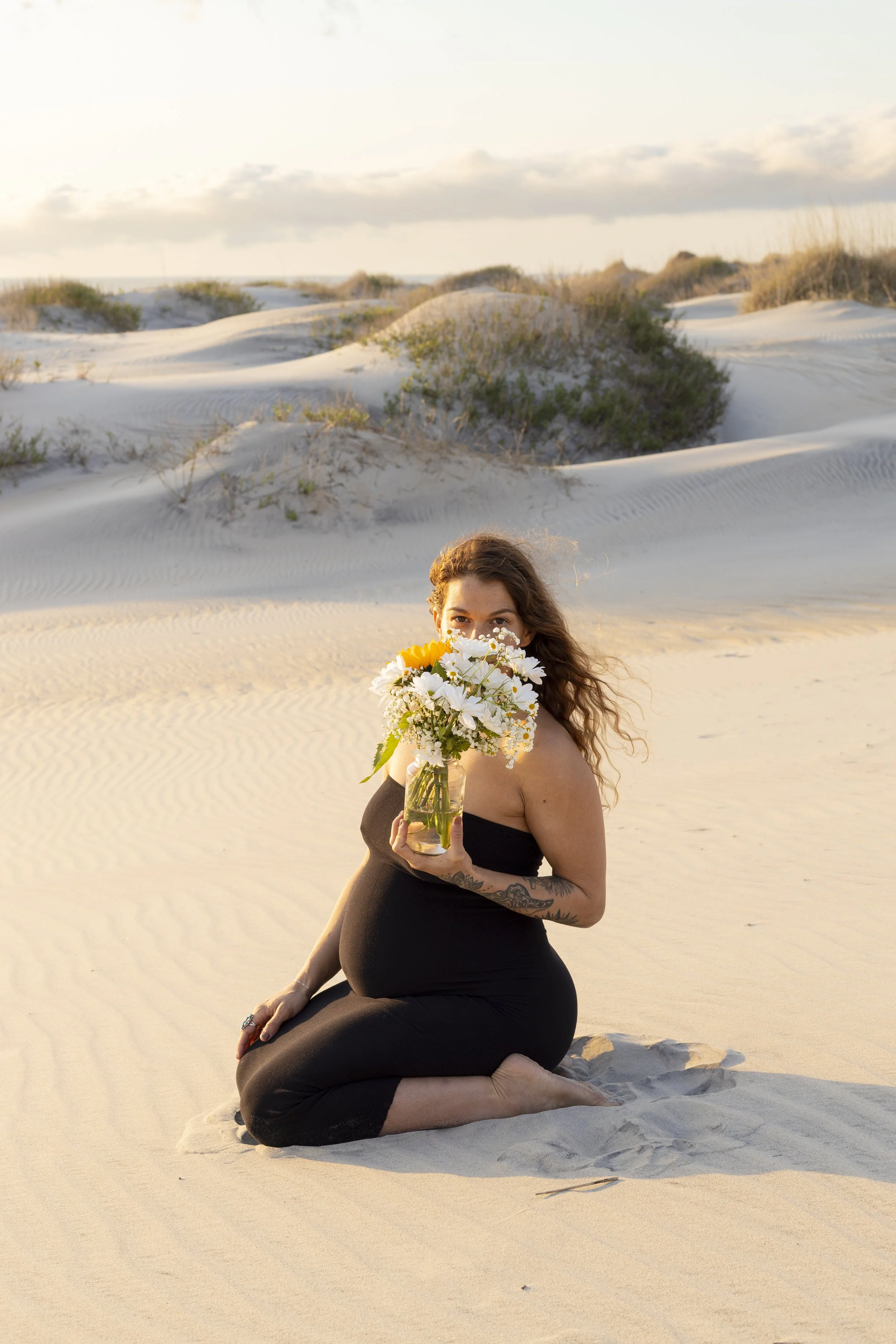 A pregnant woman kneeling in the sand at the beach, holding a bouquet of white daisies and sunflowers, with dunes and sparse vegetation in the background.