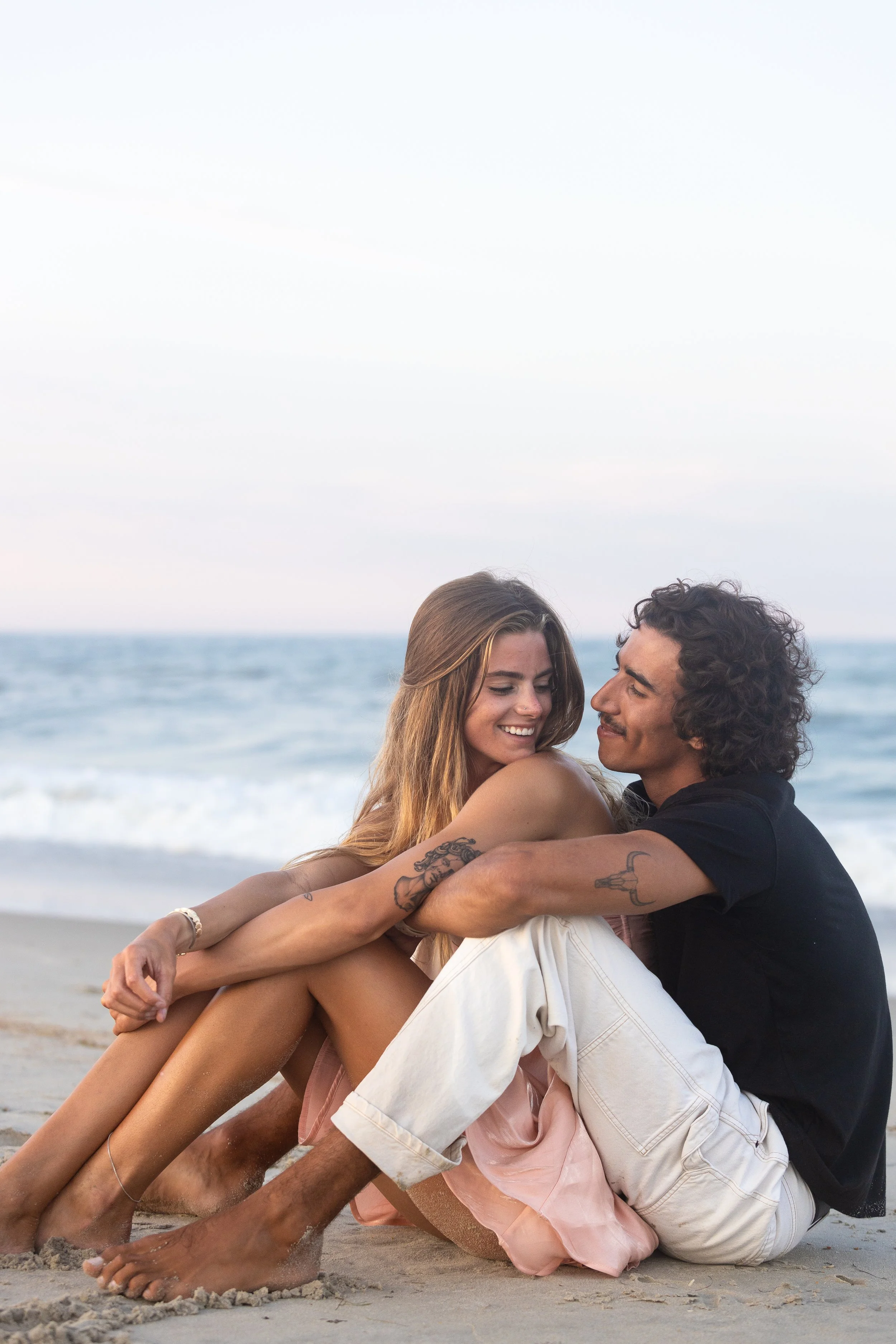 A young couple sitting on the beach, embracing and smiling at each other, with the ocean in the background.