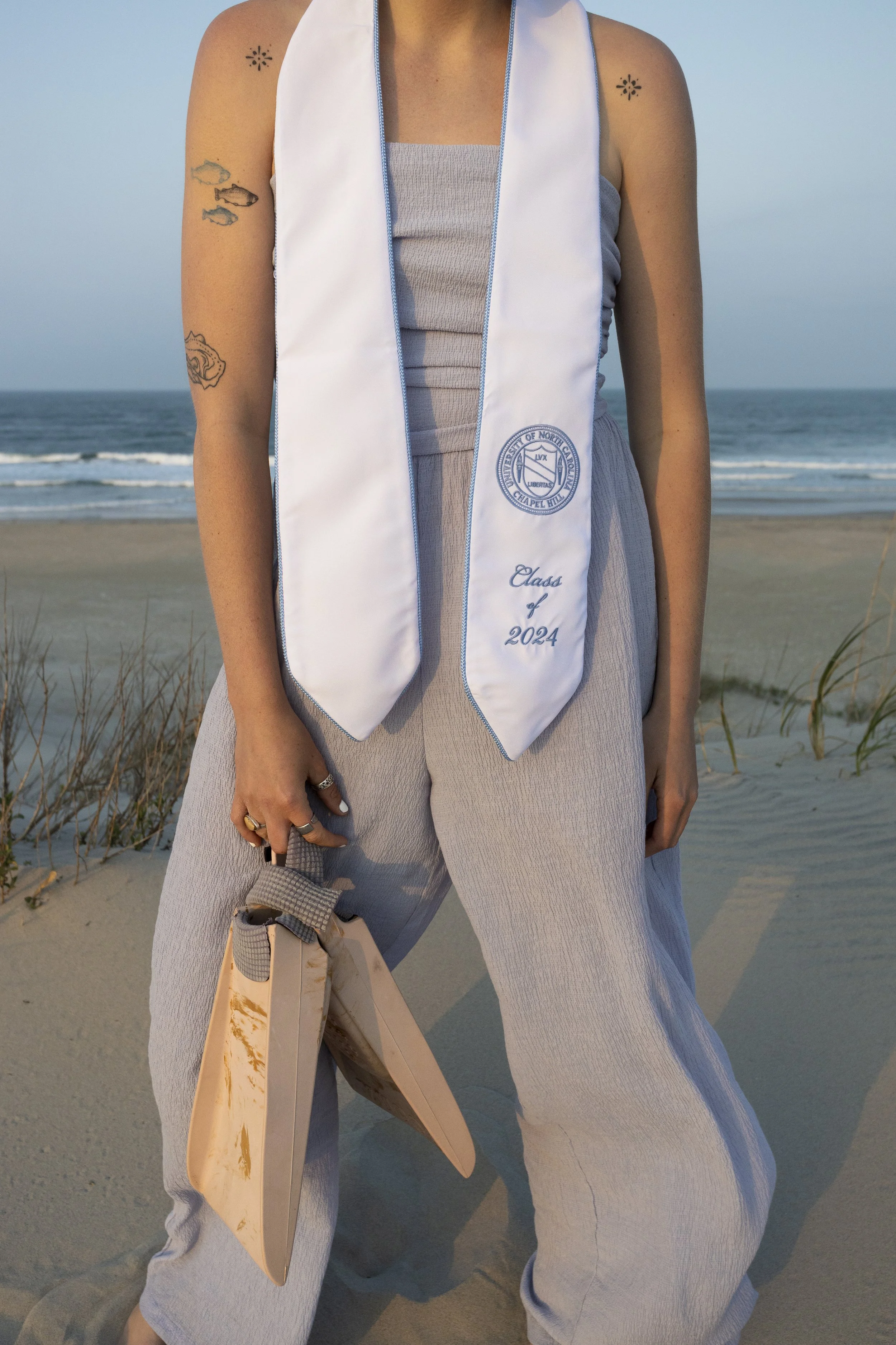 A person standing on a beach wearing a white graduation stole from the University of North Carolina at Chapel Hill, with 'Class of 2024' embroidered on it. They are holding a beige clutch purse and are dressed in light-colored clothing, with some tat