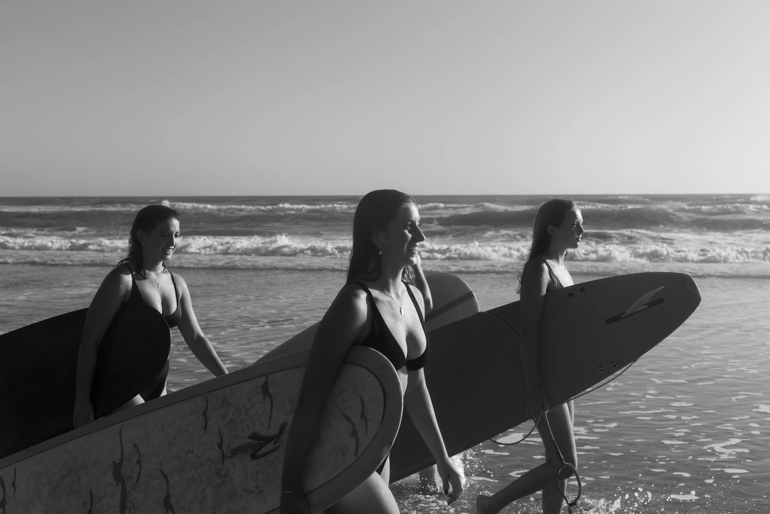 Three women in swimsuits carrying surfboards walking into the ocean at the beach during the daytime