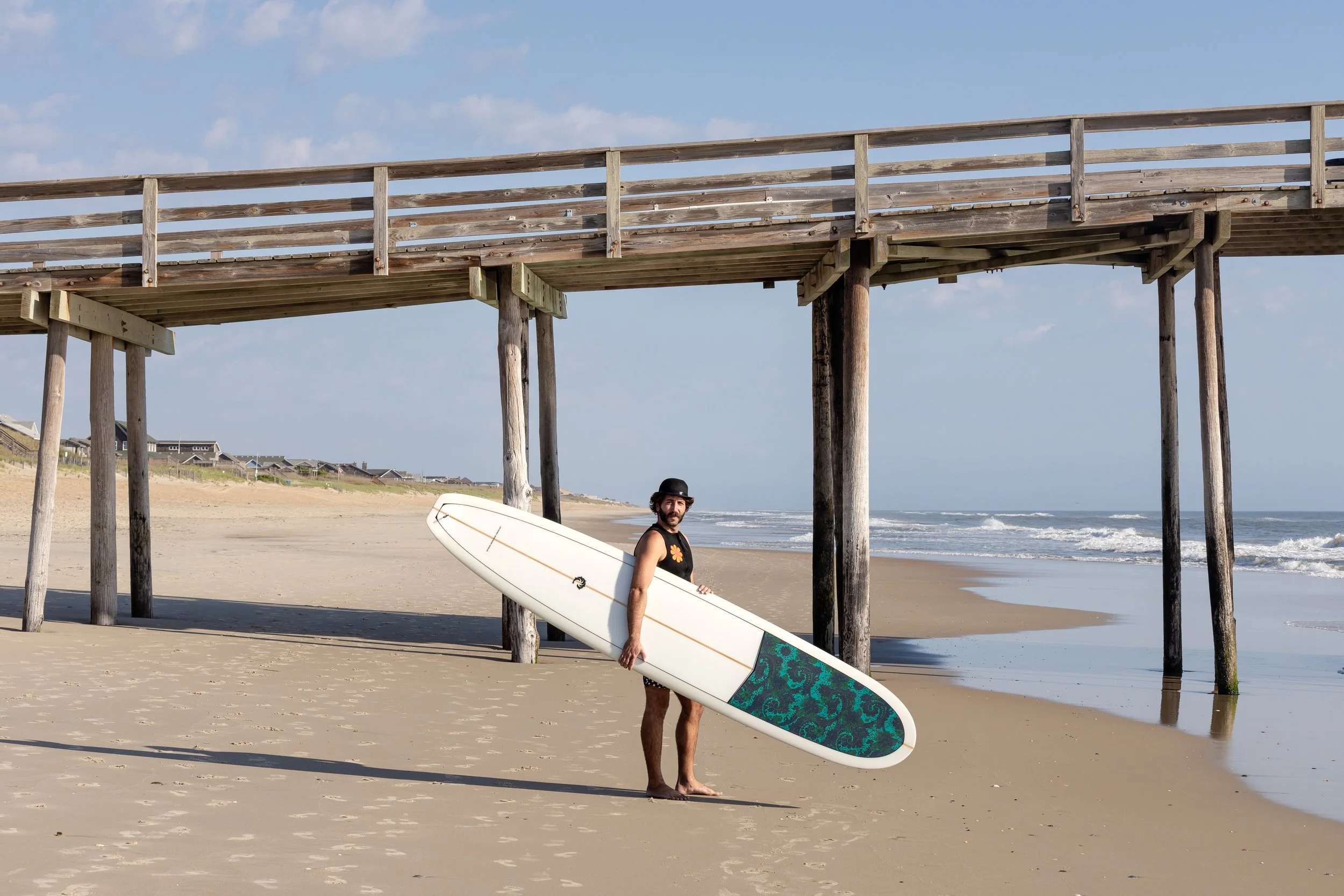 A man standing on the beach holding a surfboard under his arm, under a wooden pier, with the ocean and waves in the background.