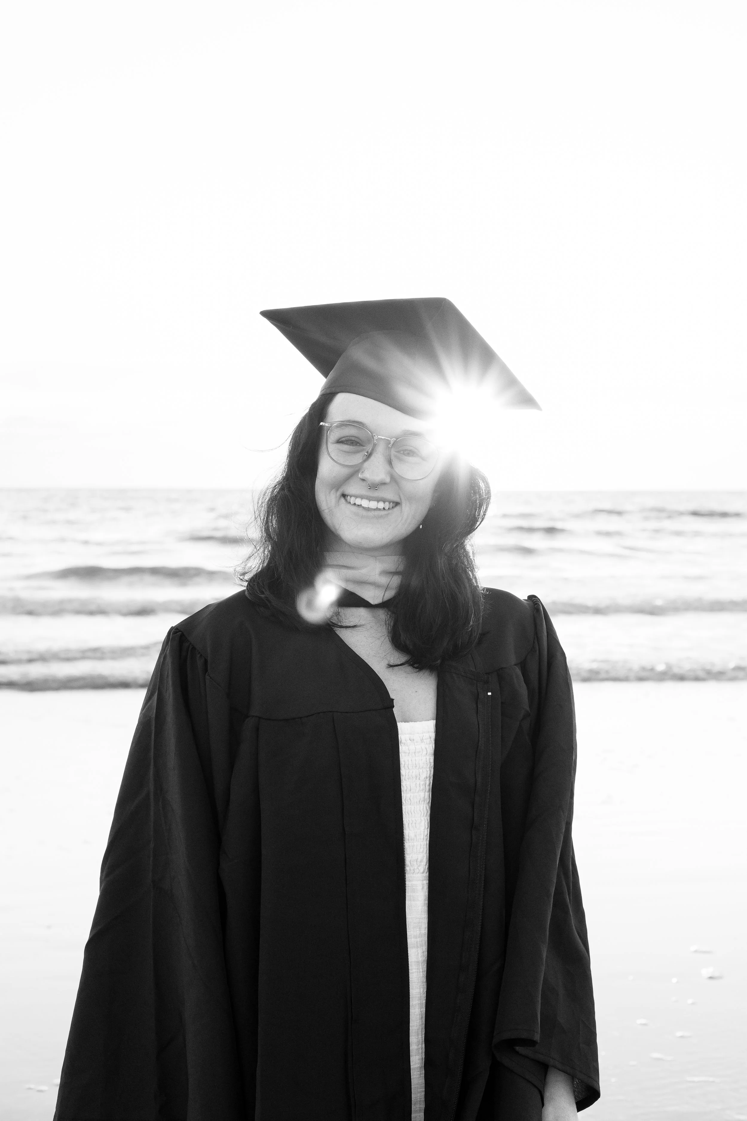 A woman wearing graduation gown and cap, smiling on the beach during sunset, captured in black and white.
