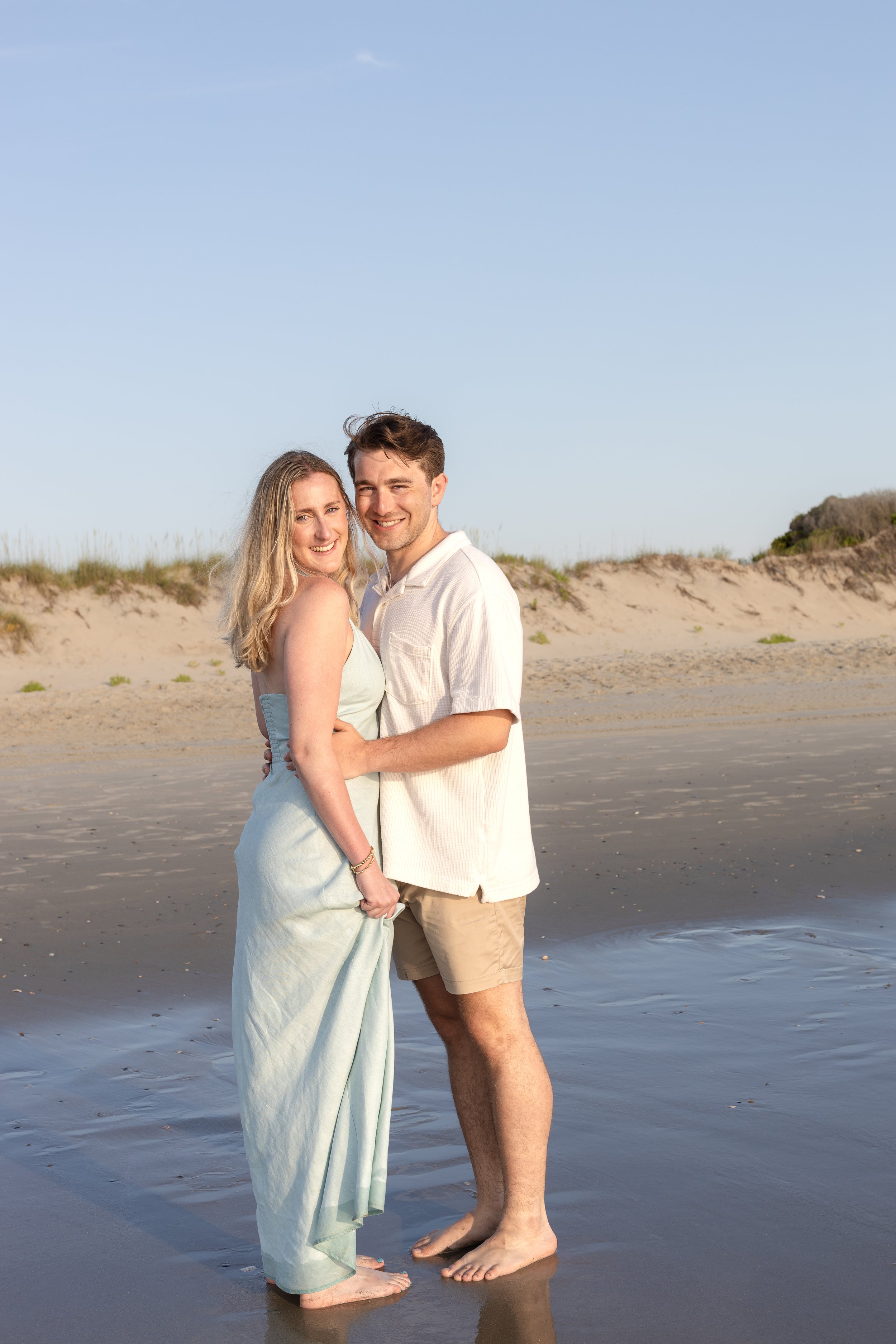 A young couple stands in shallow water on a beach, smiling and embracing each other.