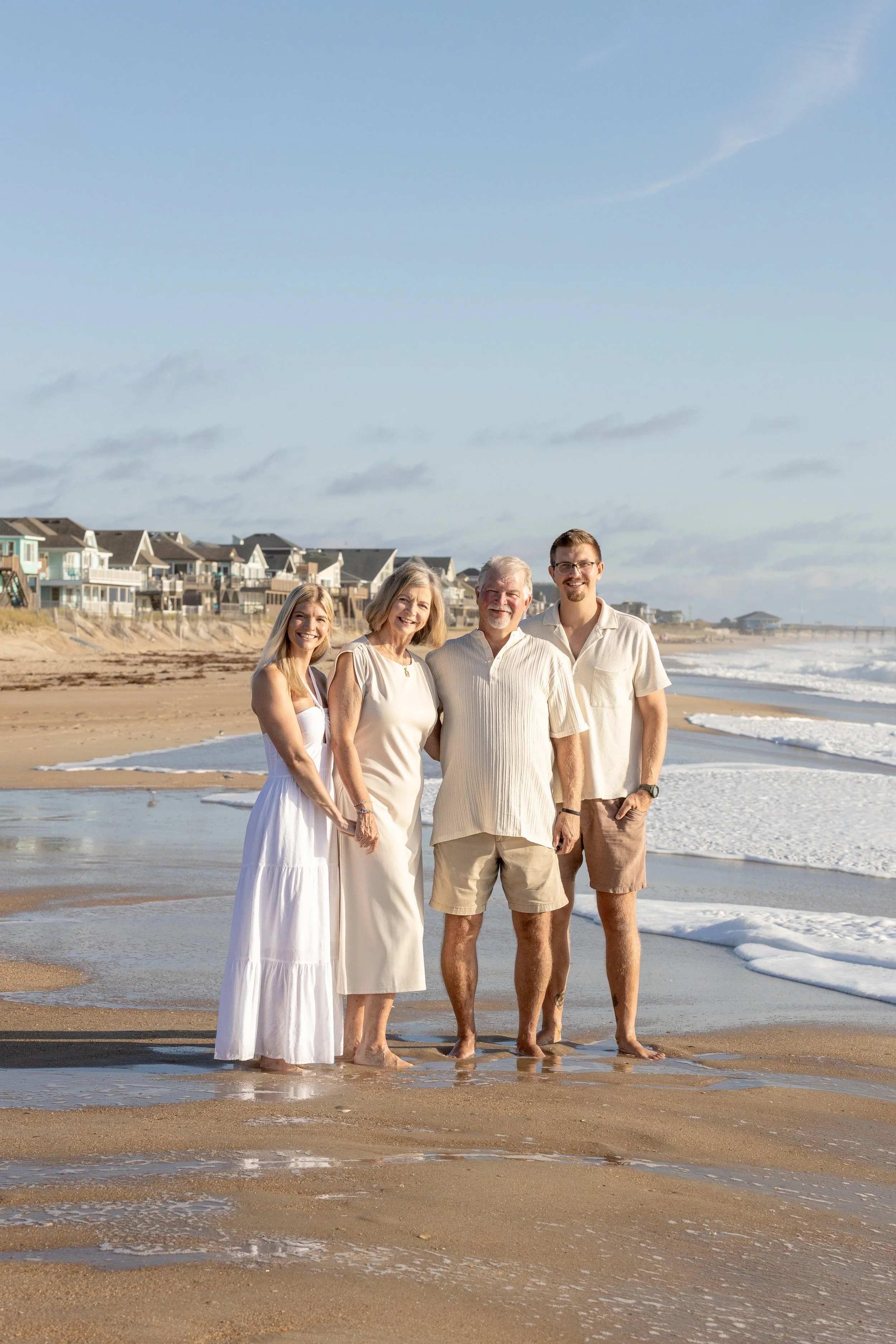 A family of four standing on a beach near the shoreline with houses in the background, smiling at the camera.
