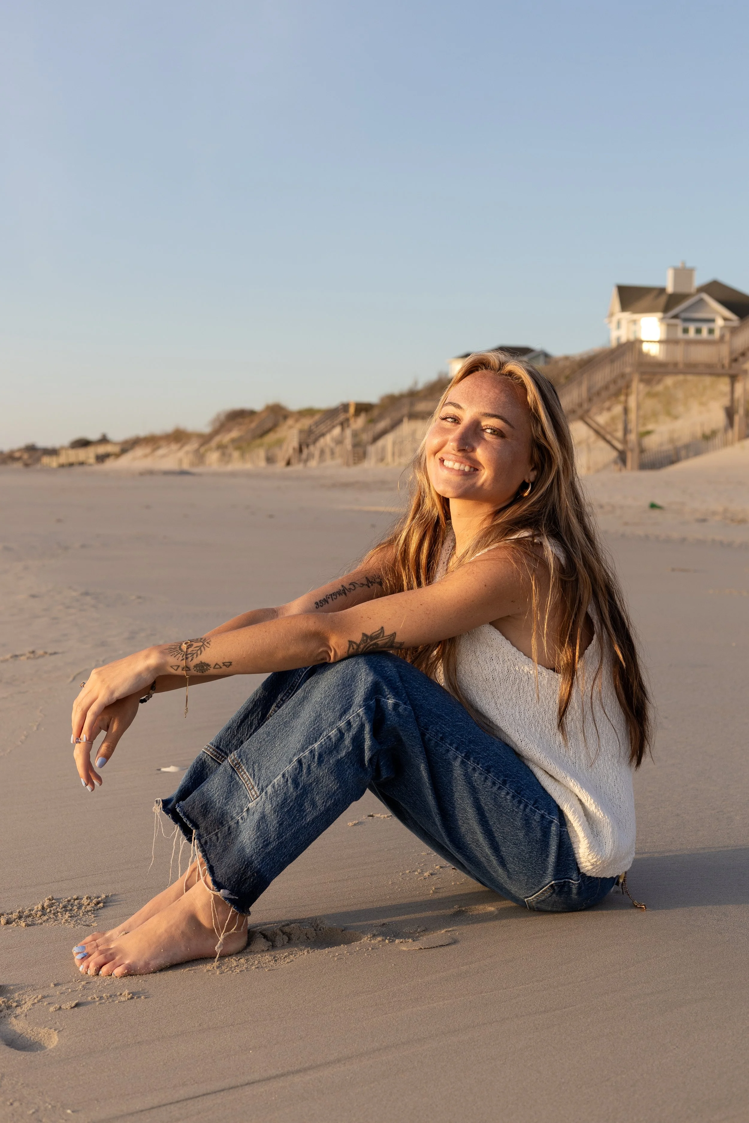 A smiling young woman with long hair sitting on a sandy beach during sunset, with houses on stilts in the background.