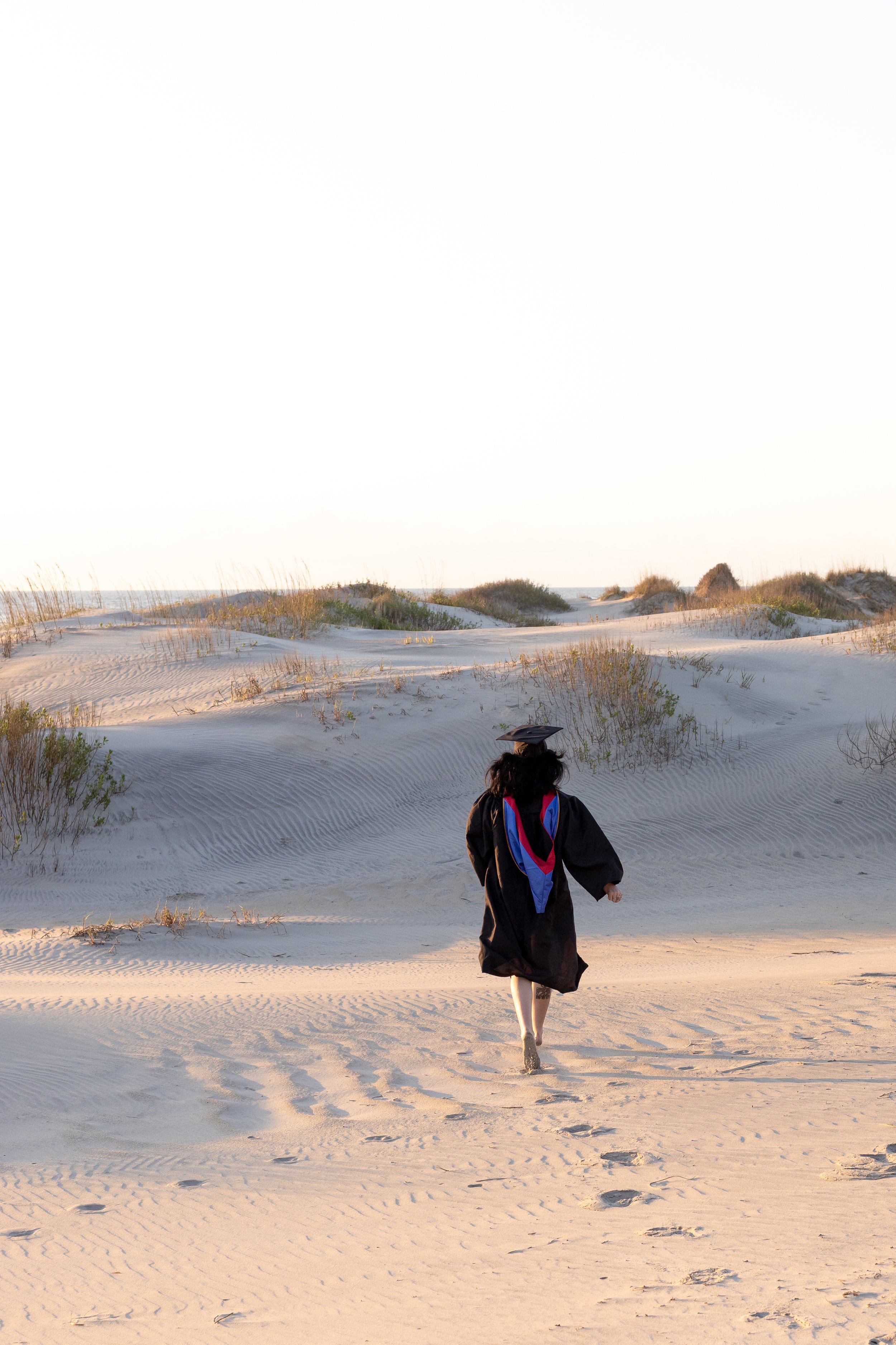 A person in a graduation cap and gown walking on a sandy beach with sparse vegetation, heading towards dunes and an overcast sky.