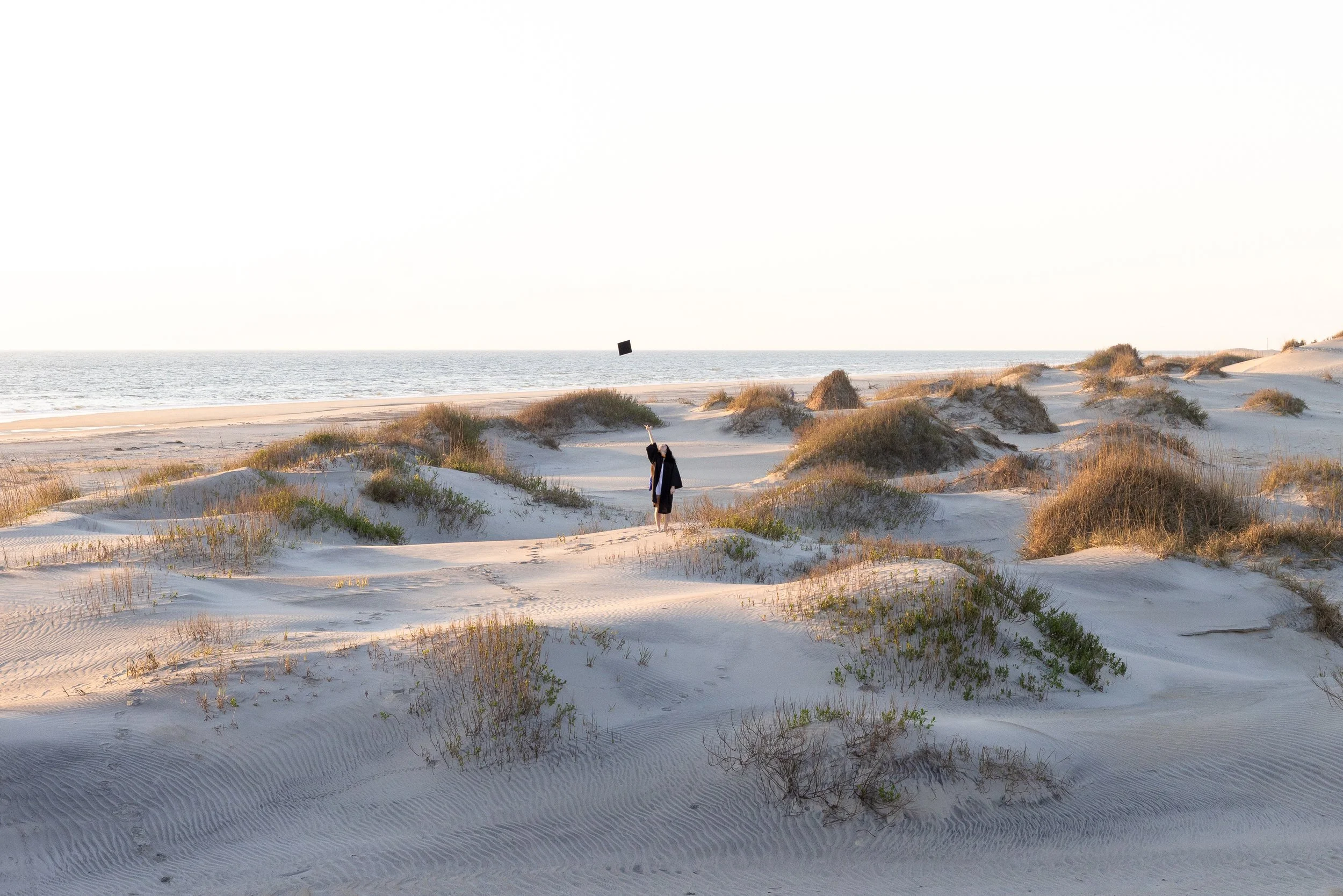 A person standing on sandy dunes near the ocean, tossing a hat into the air.