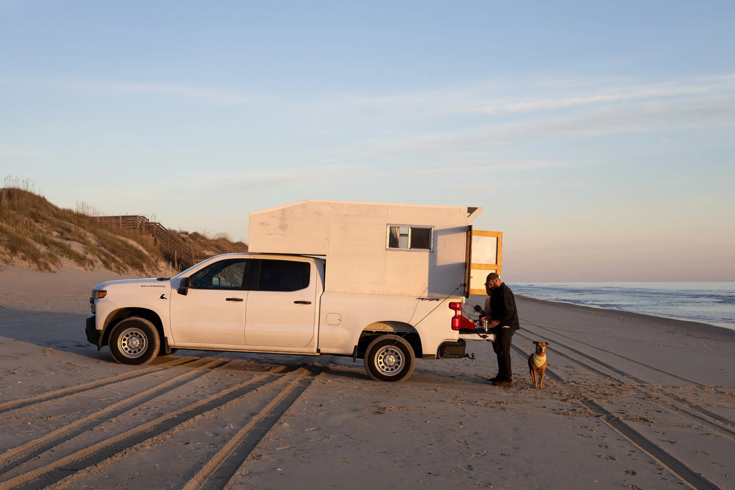 A white pickup truck with a homemade camper parked on a sandy beach, with a man and a dog next to it during sunset.