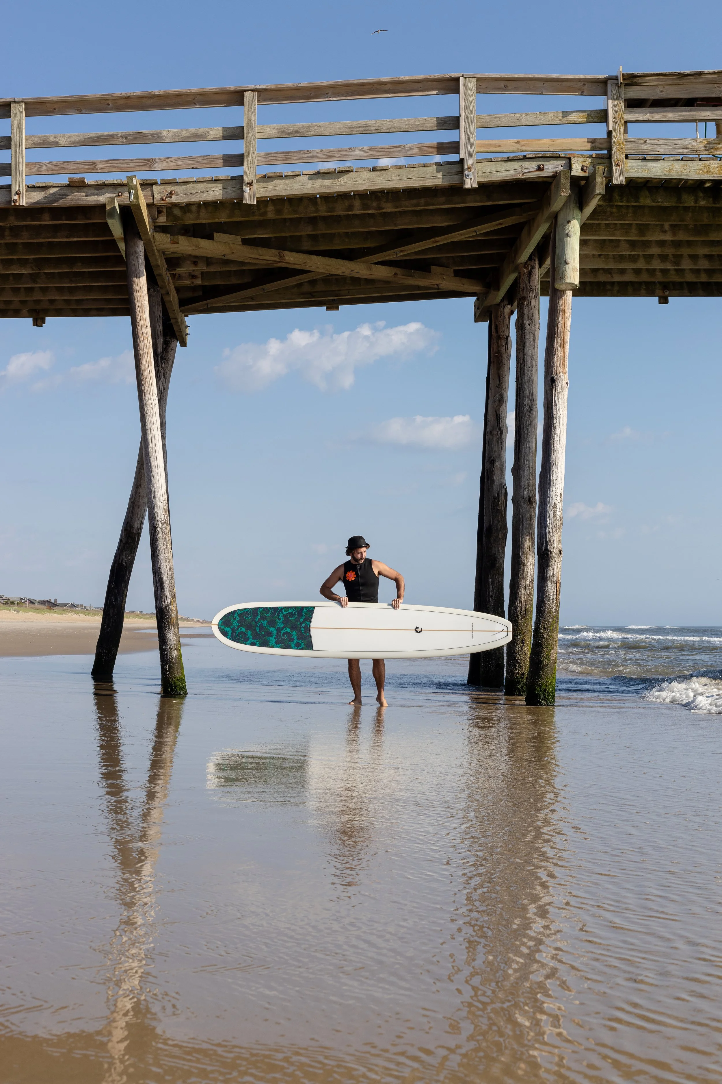 A man holding a surfboard under a wooden pier at the beach, with a blue sky and ocean in the background.
