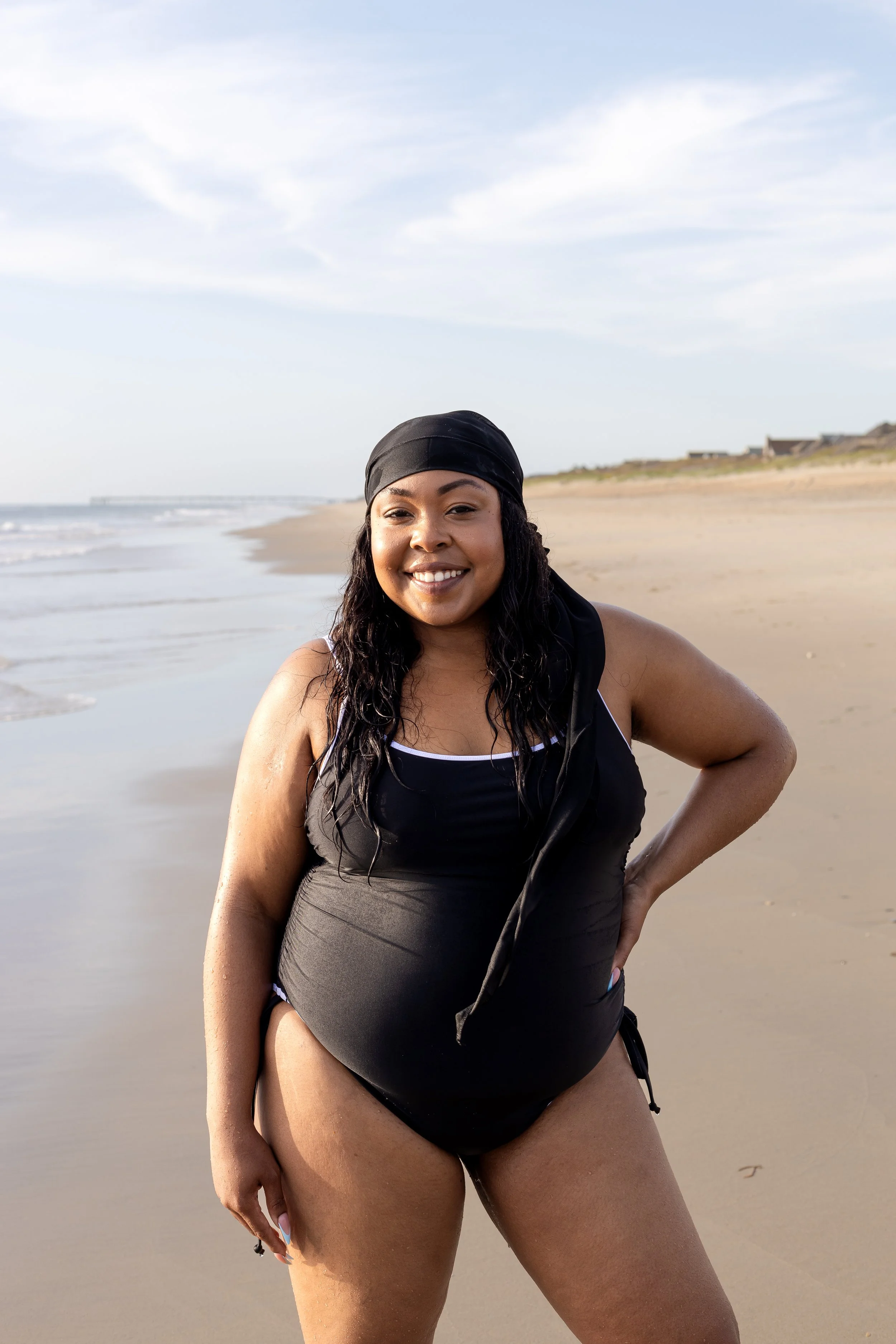 A woman in a black swimsuit and black headscarf smiling at the camera on a beach with sand and ocean waves in the background.
