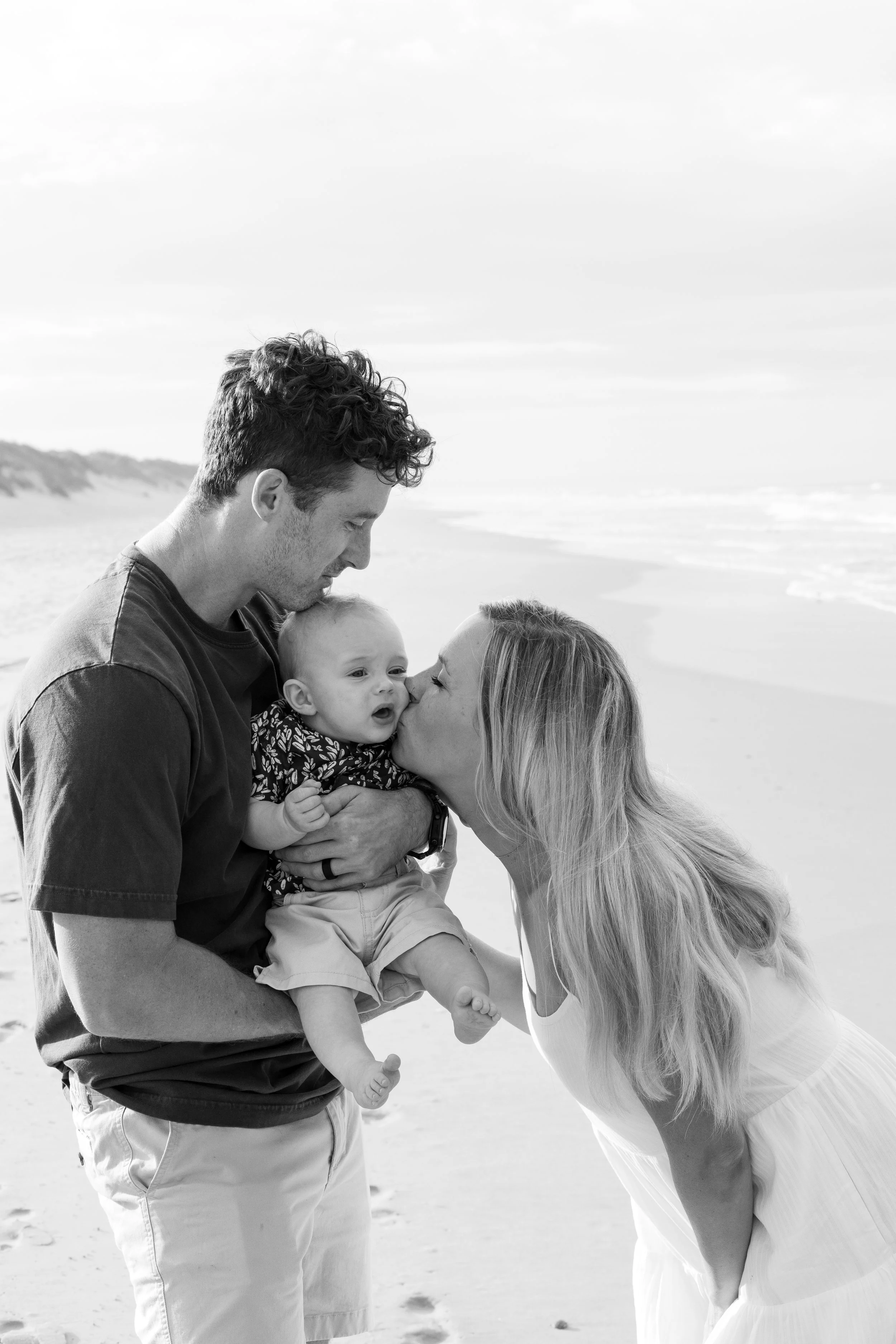 A black and white photo of a family at the beach, with a man holding a toddler and a woman giving a kiss to the toddler, all sharing a tender moment.