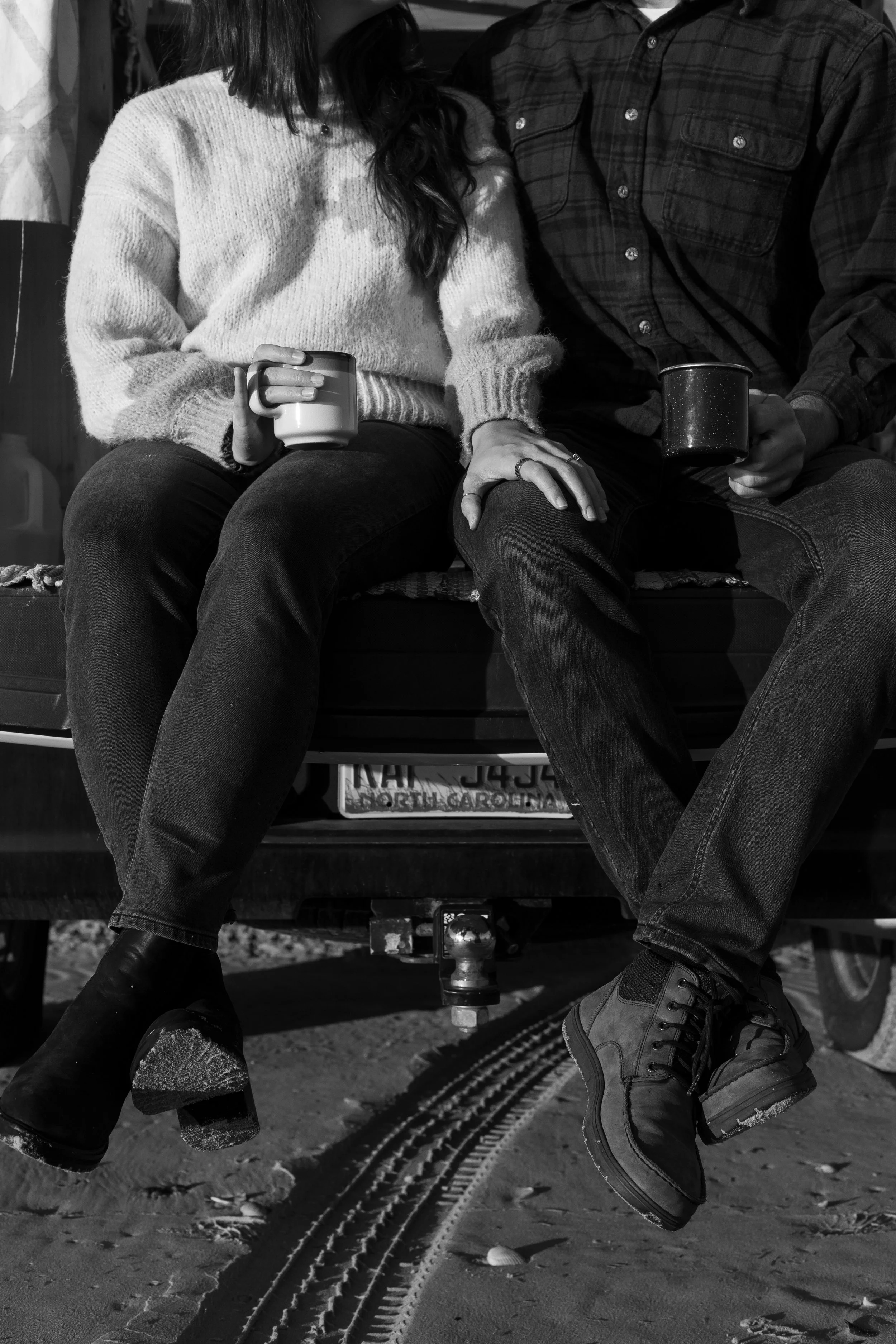 A black and white photo of a couple sitting on the back of a truck, holding mugs, with their legs hanging over the edge, at the beach or sandy area.