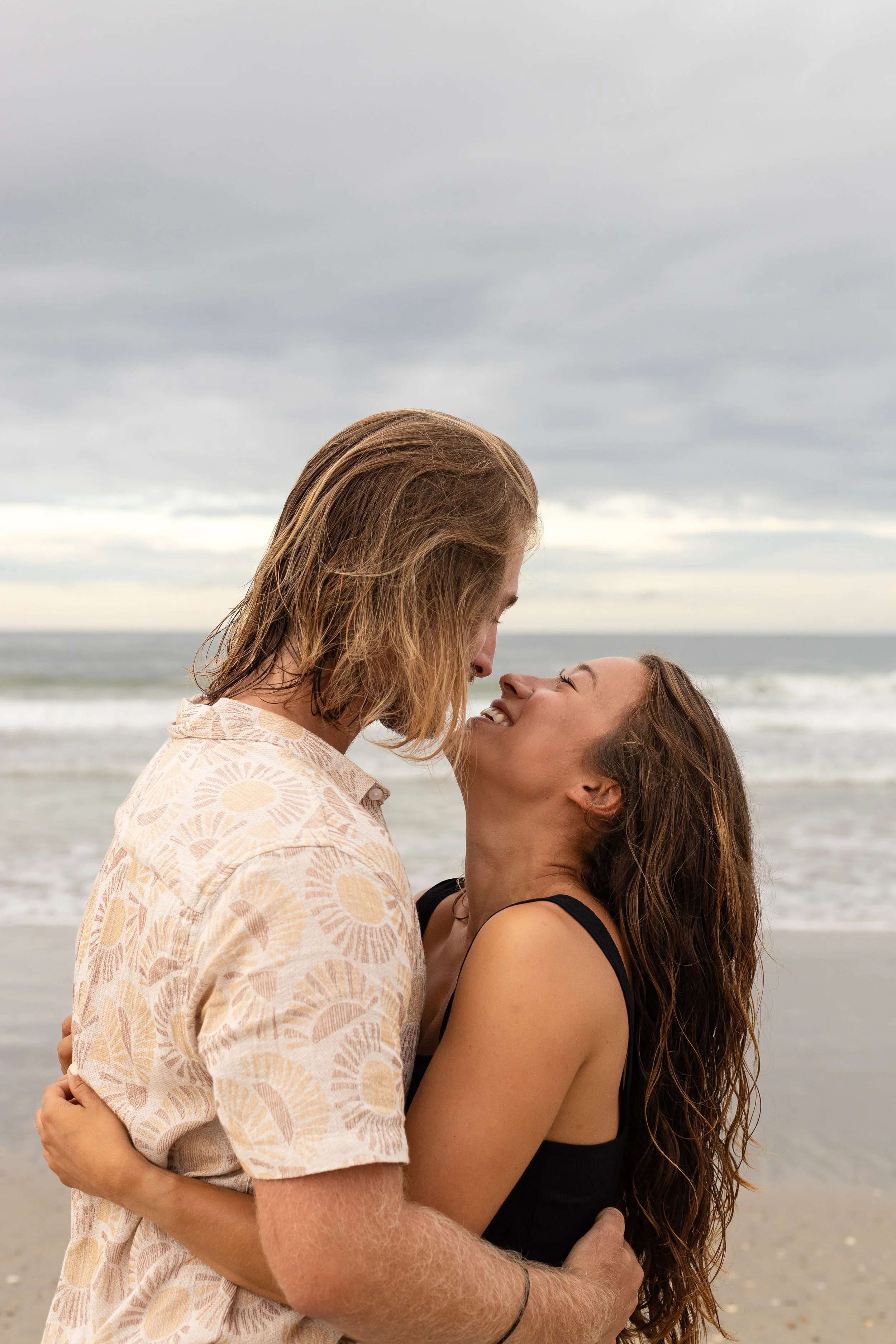 A couple standing close on the beach, smiling and looking into each other's eyes, with ocean waves and a cloudy sky in the background.