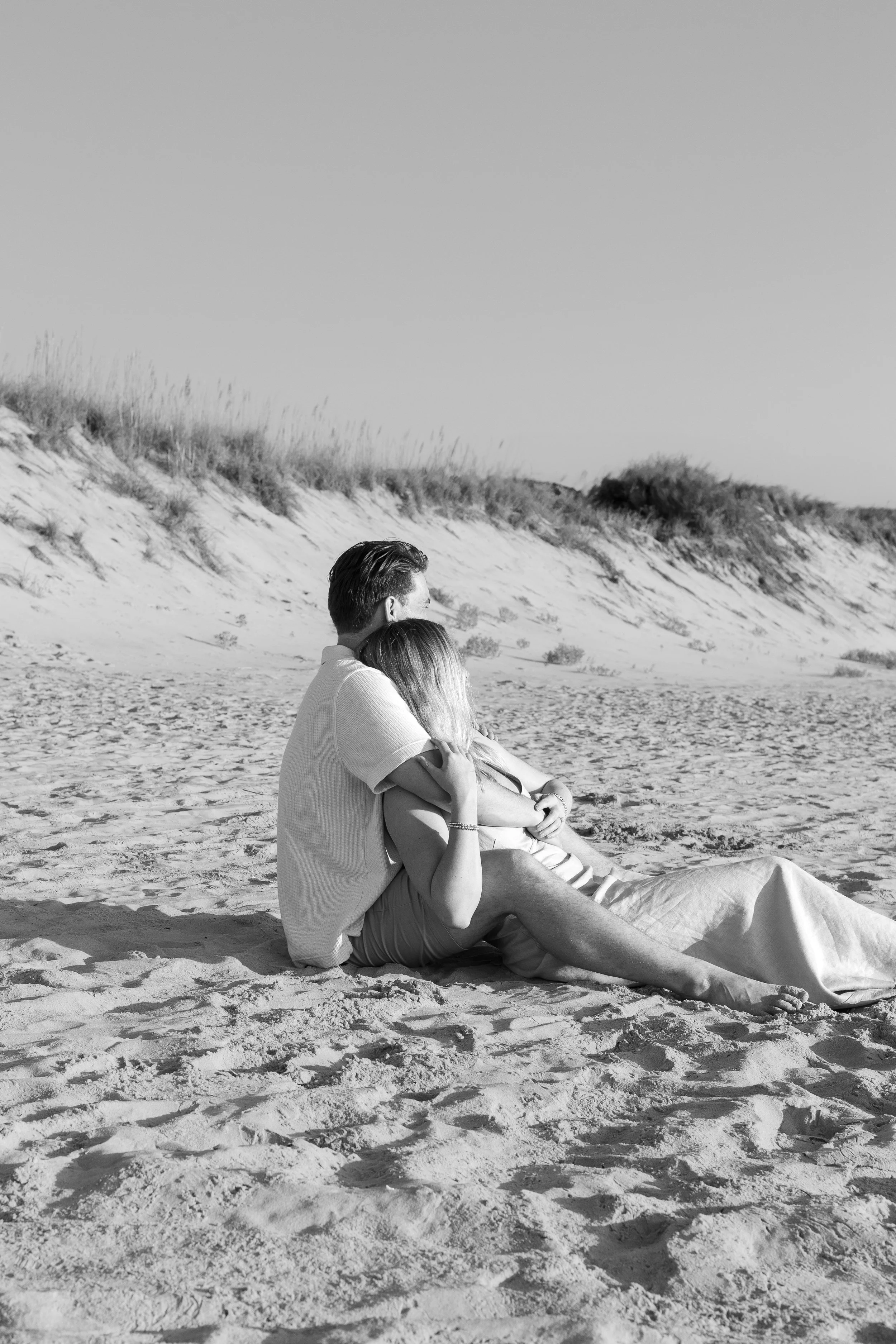 A black-and-white photo of a couple sitting on a sandy beach, embracing each other and looking towards the horizon, with dune grass and sand dunes in the background.