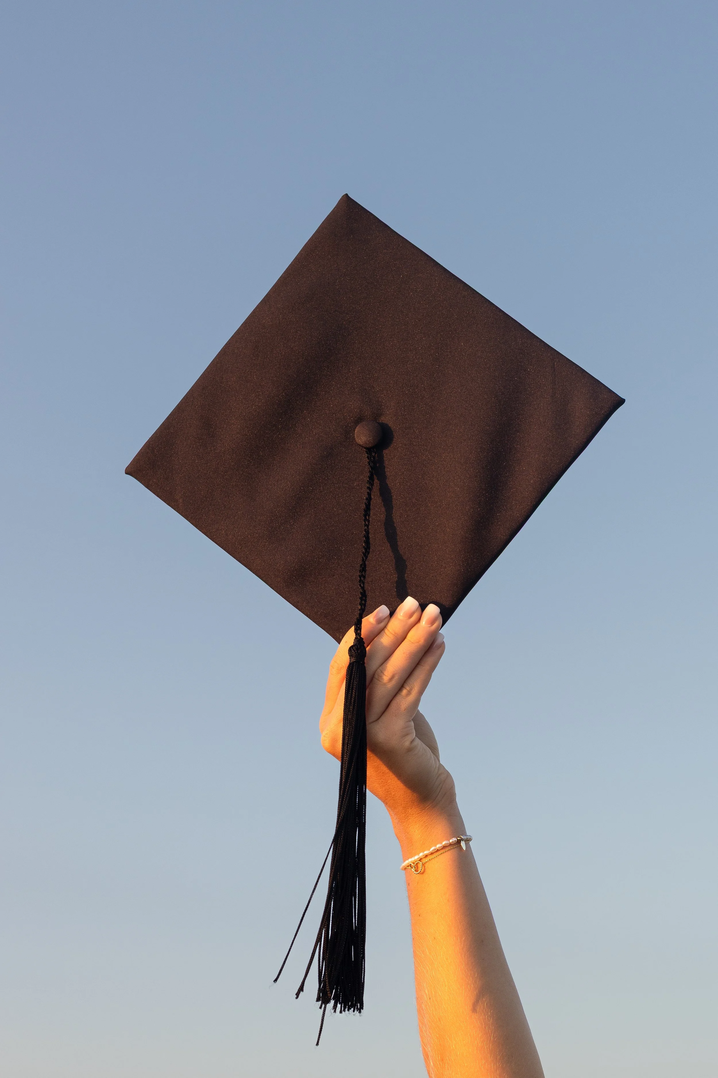 A hand holding a black graduation cap against a blue sky.
