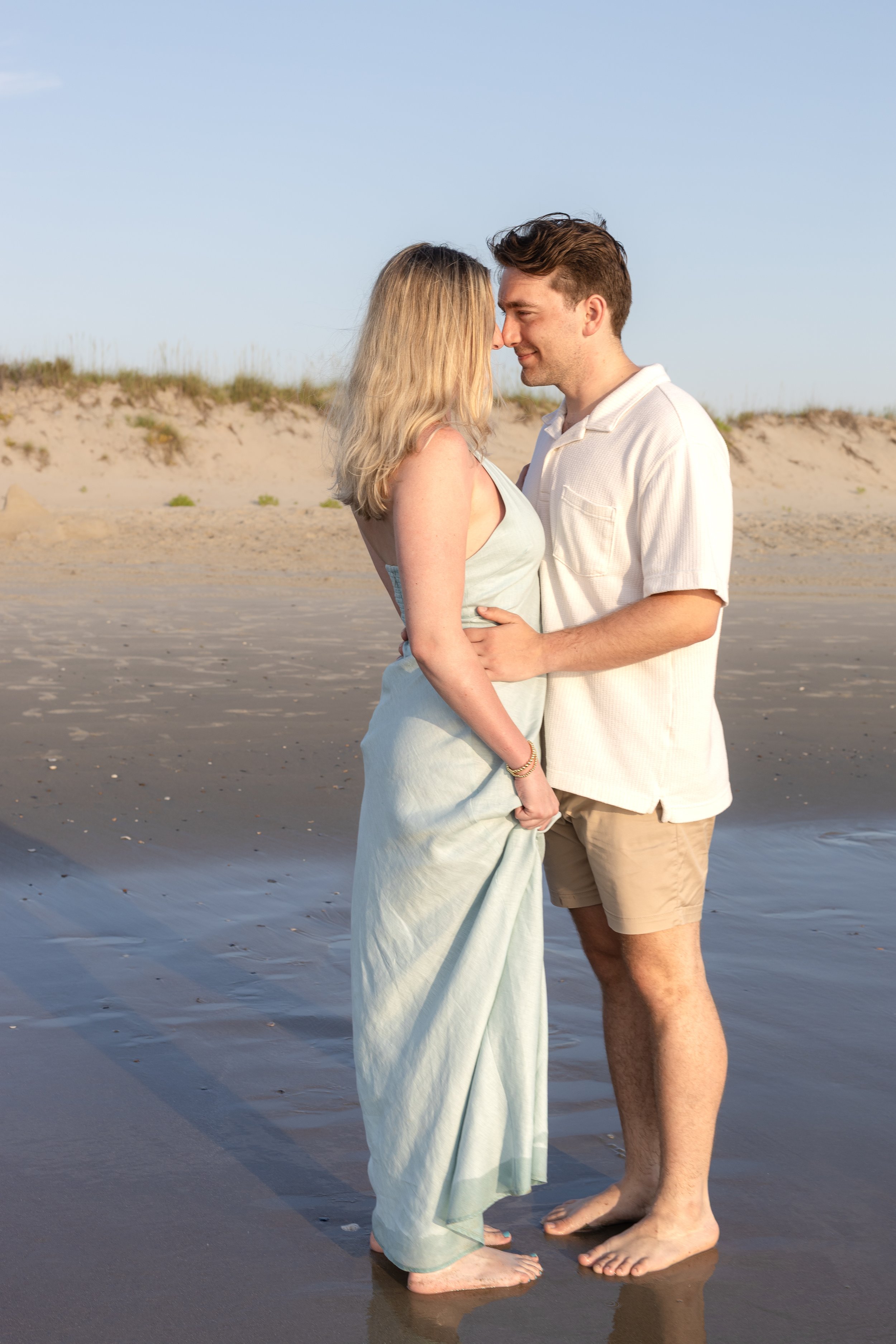 A couple standing close on the beach, smiling and touching foreheads, with ocean and sandy dunes in the background.