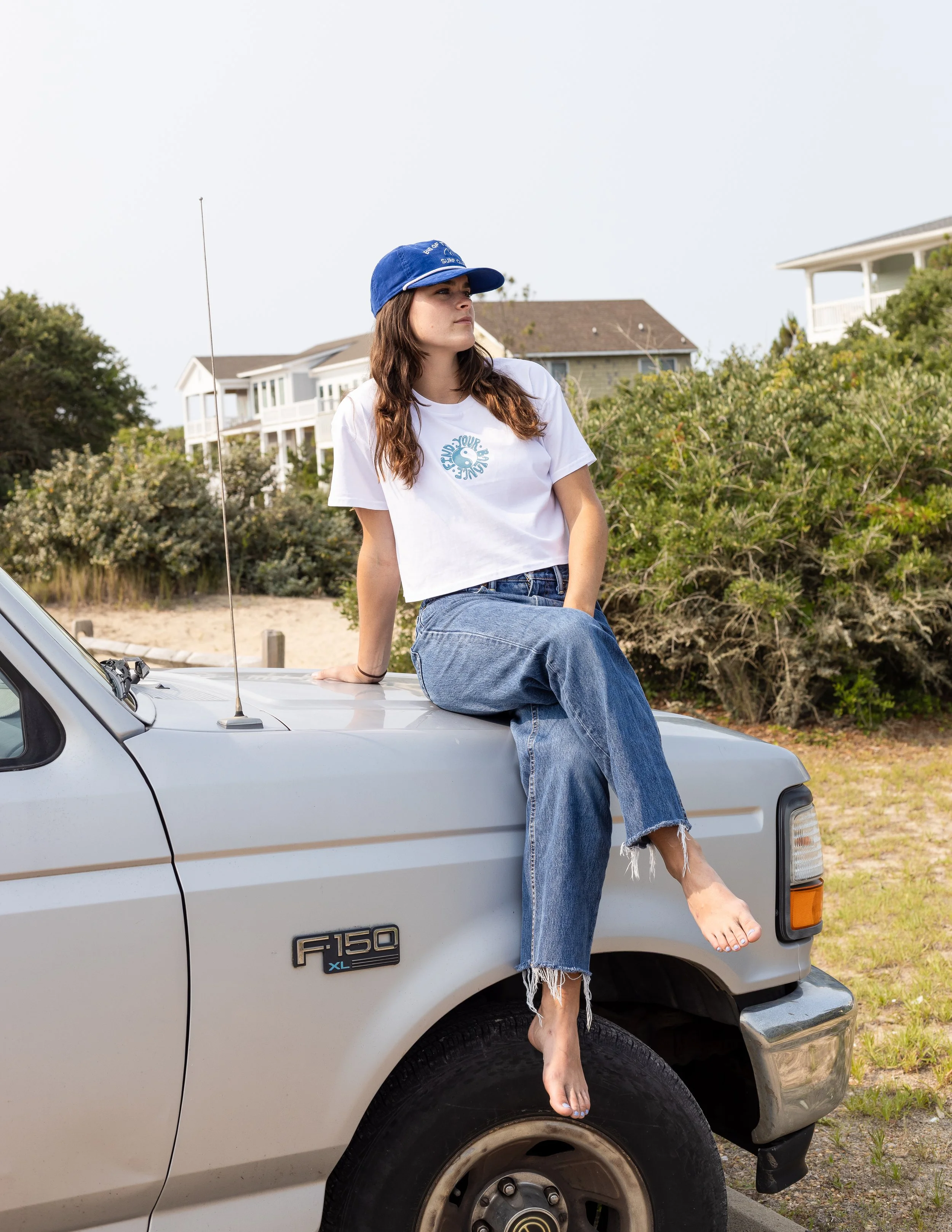 A young woman with long brown hair, wearing a white T-shirt, blue jeans, and a blue cap, sitting on the hood of a gray Ford F150 truck in an outdoor area near houses and bushes.