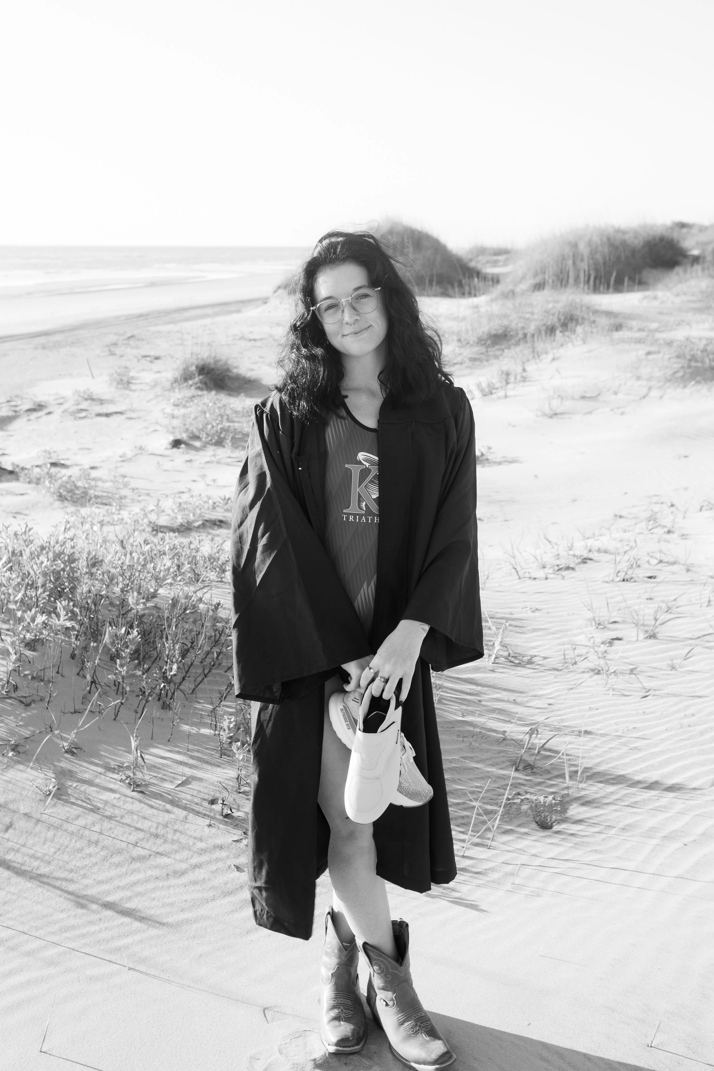 A young woman standing on a sandy beach holding a pair of running shoes, wearing boots, a triathlon shirt, and a graduation gown, with dunes and ocean in the background.