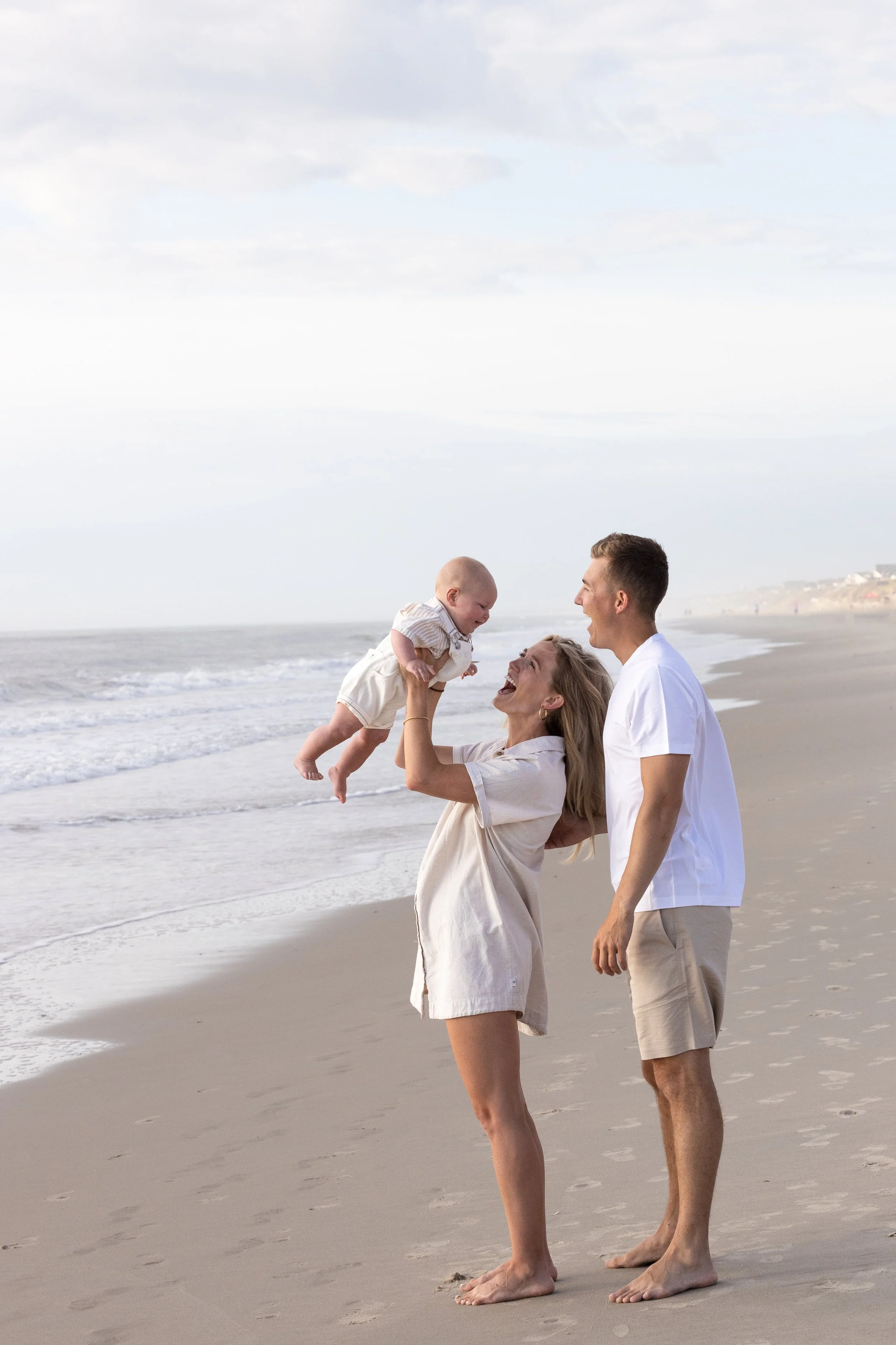 A family of three playing on a beach with waves in the background, with the mother lifting a baby in her hands while the father stands nearby, smiling at them.