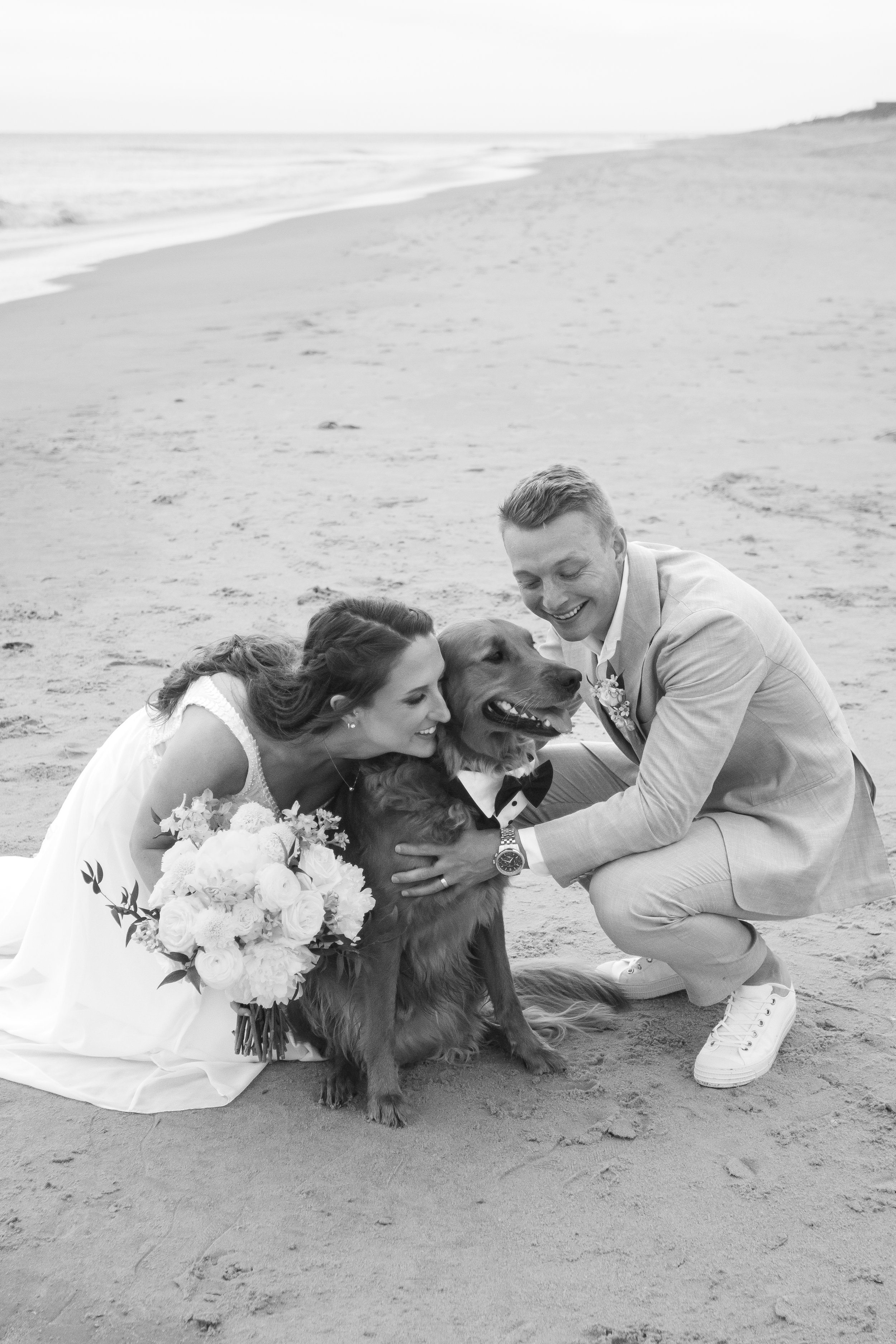 A bride and groom on the beach, kneeling next to their dog dressed in a tuxedo, all smiling and happy, with the bride holding a bouquet of flowers.