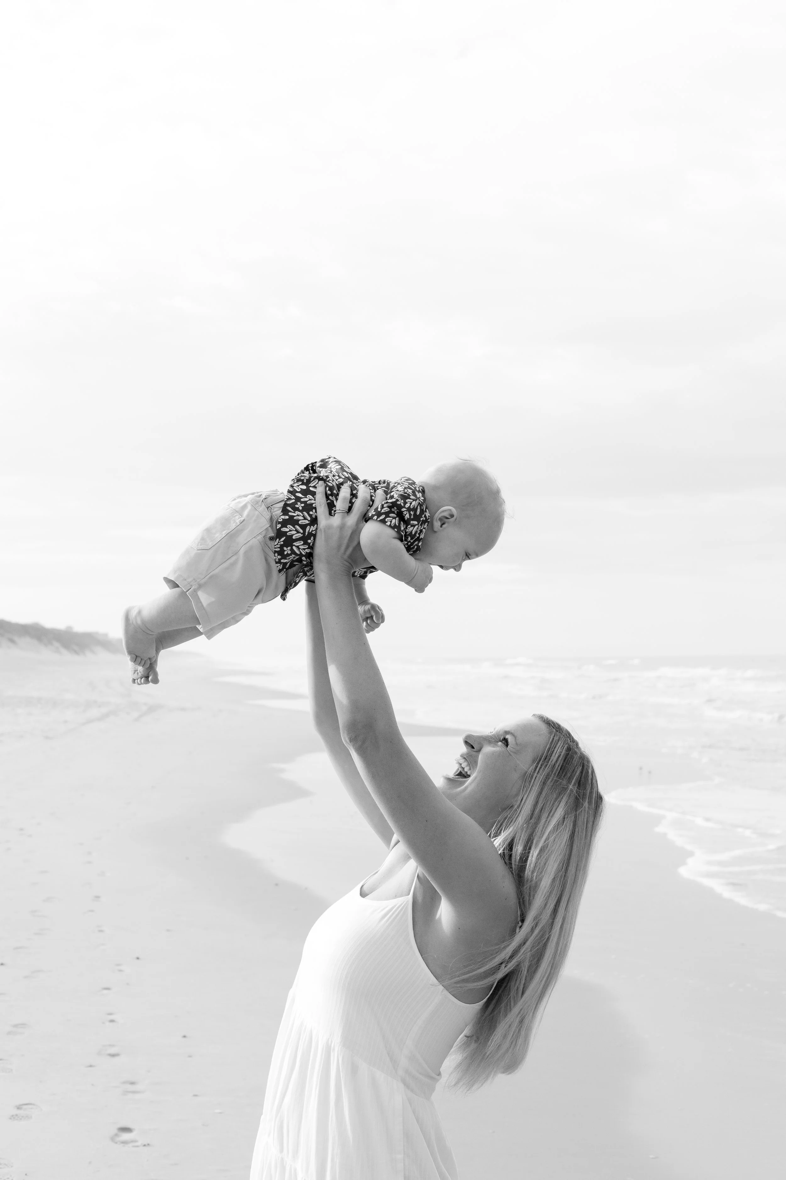 A woman at the beach lifting her baby in the air, both smiling and enjoying the moment.