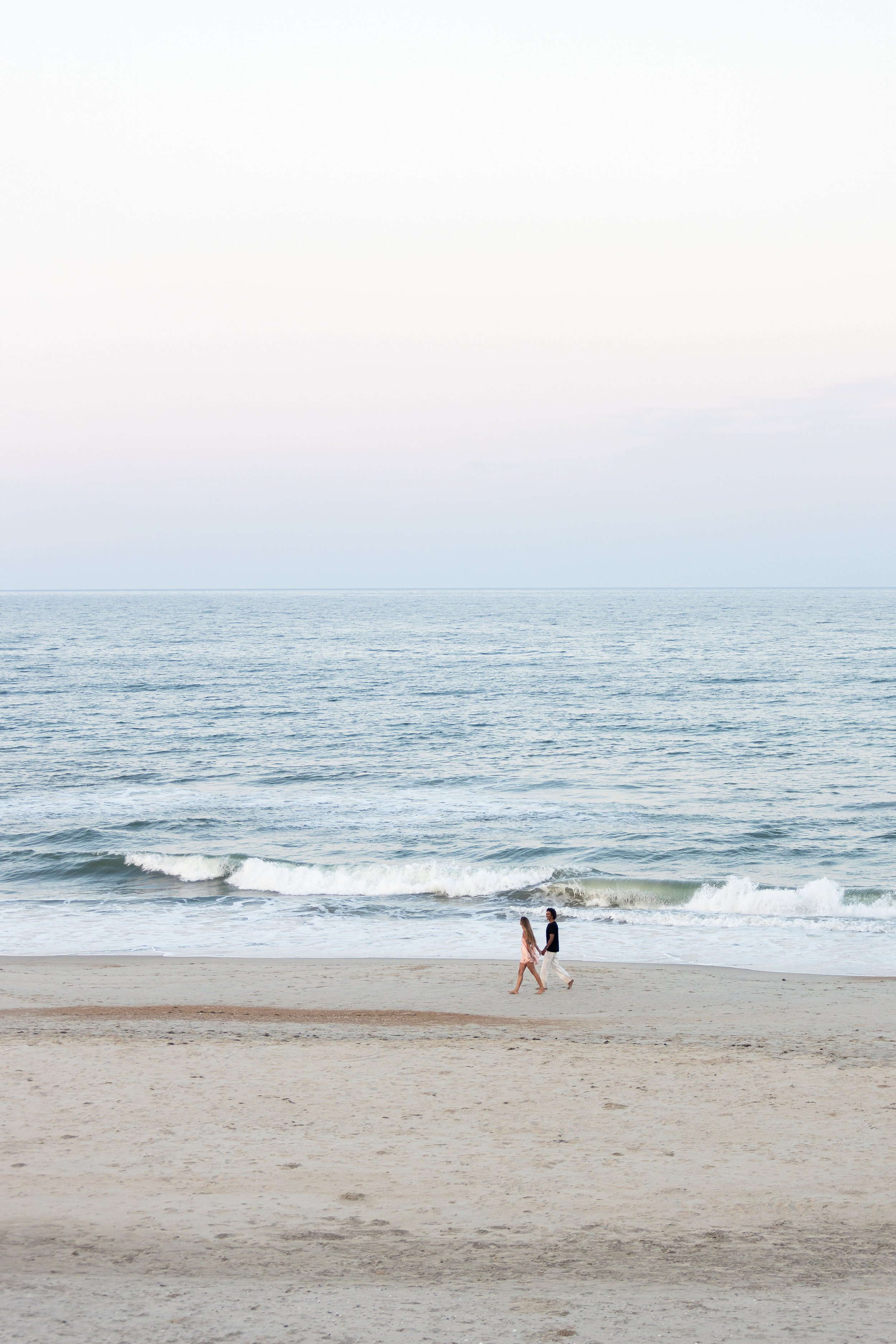 A couple walking along the beach near the shoreline with gentle waves in the ocean, under a mostly clear sky.