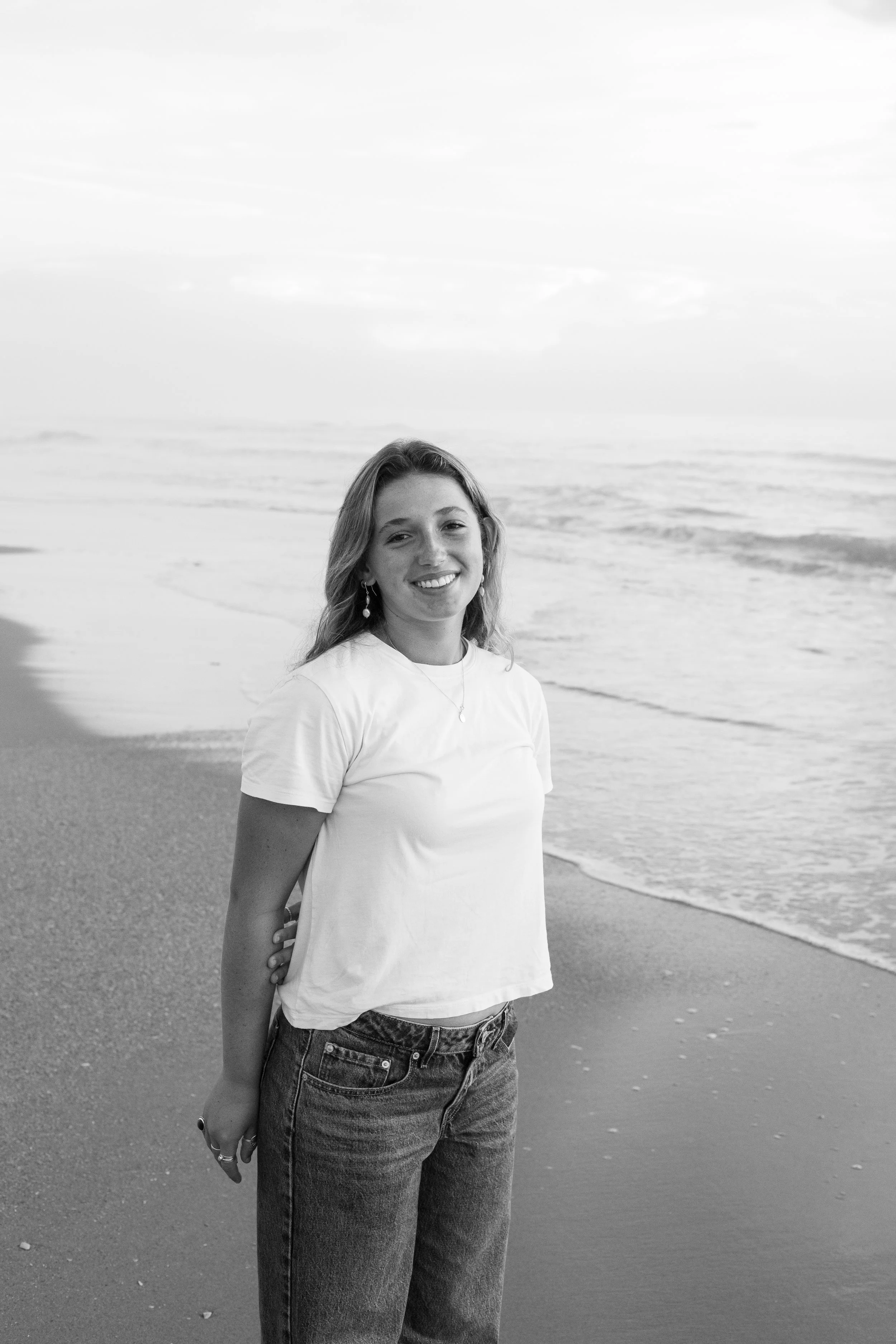 A young woman with long wavy hair, wearing a white t-shirt and jeans, smiling and standing on the beach near the ocean, in black and white.