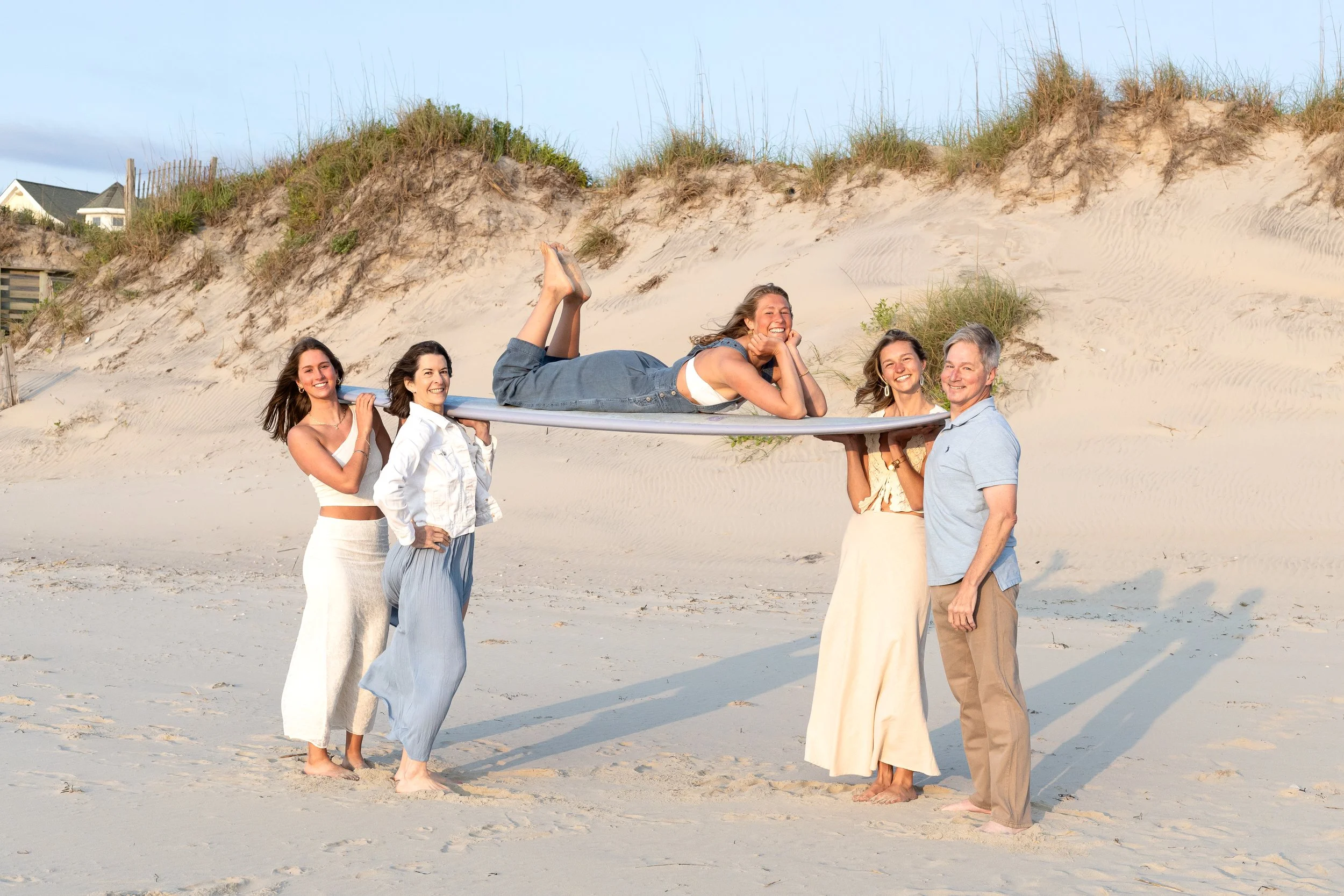 Five people on a beach, four women and one man, holding a surfboard. One woman is lying on the surfboard, smiling, while the others stand supporting it. Sand dunes and houses are in the background.