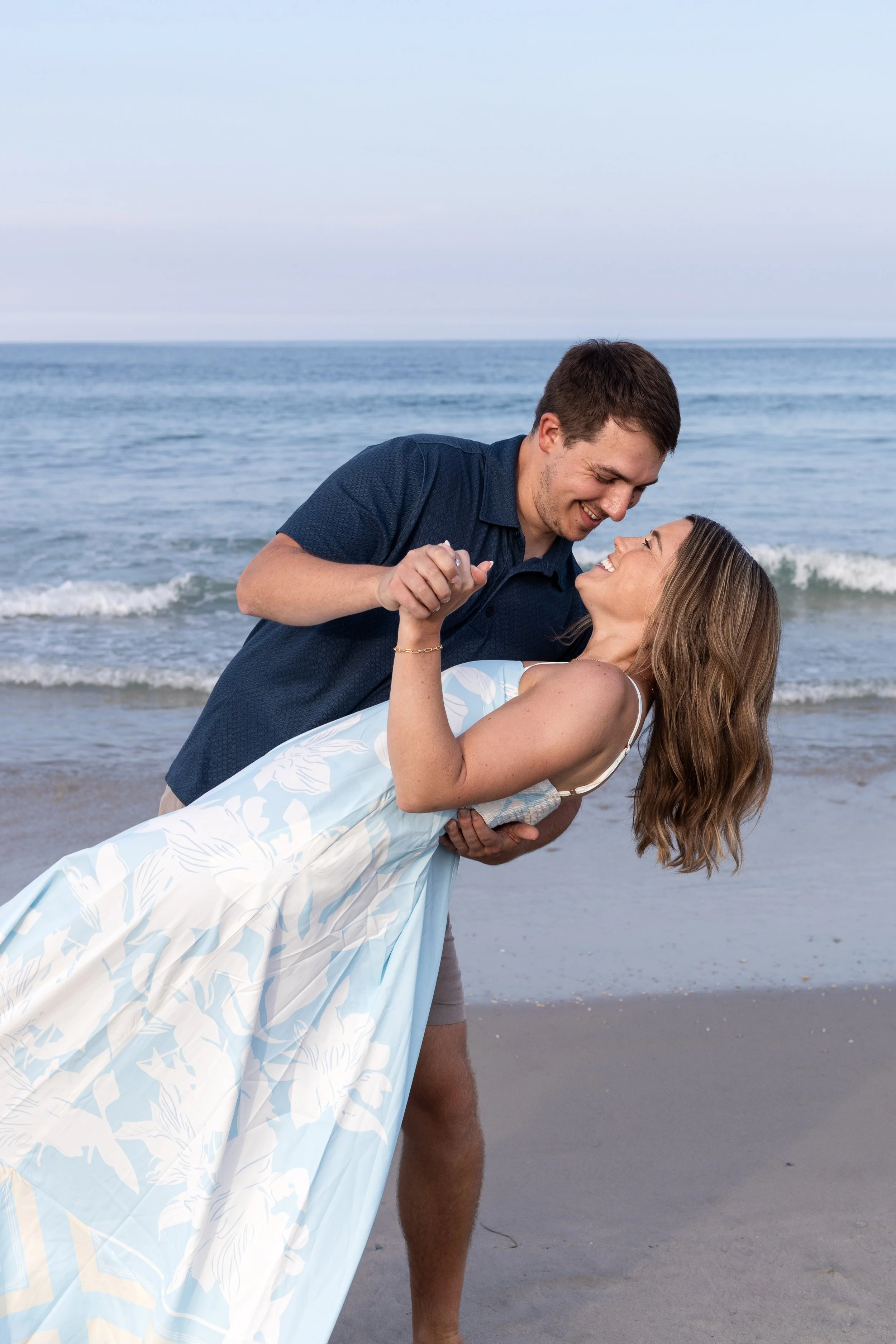 A couple dancing on the beach by the ocean, with the man dipping the woman while holding her hand, both smiling and looking at each other.