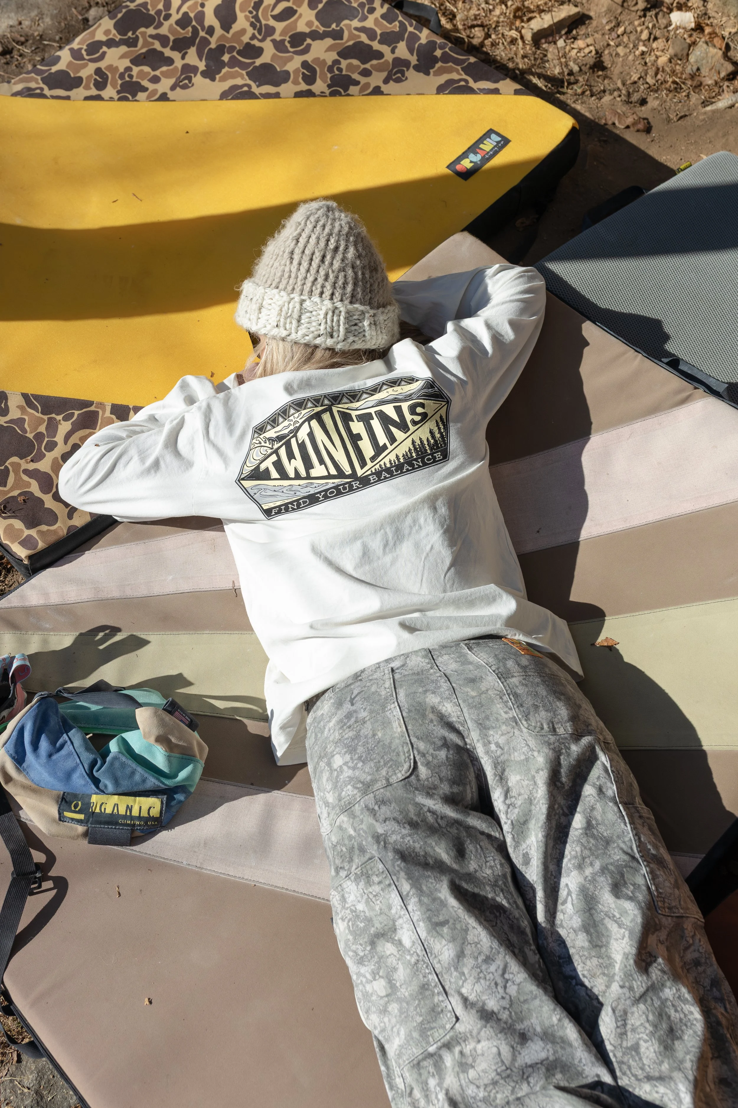 Person lying face down outdoors on a bench, wearing a beanie, a white T-shirt with 'TWIN TENS' graphic, and gray patterned pants, with camping gear and a skateboard nearby.