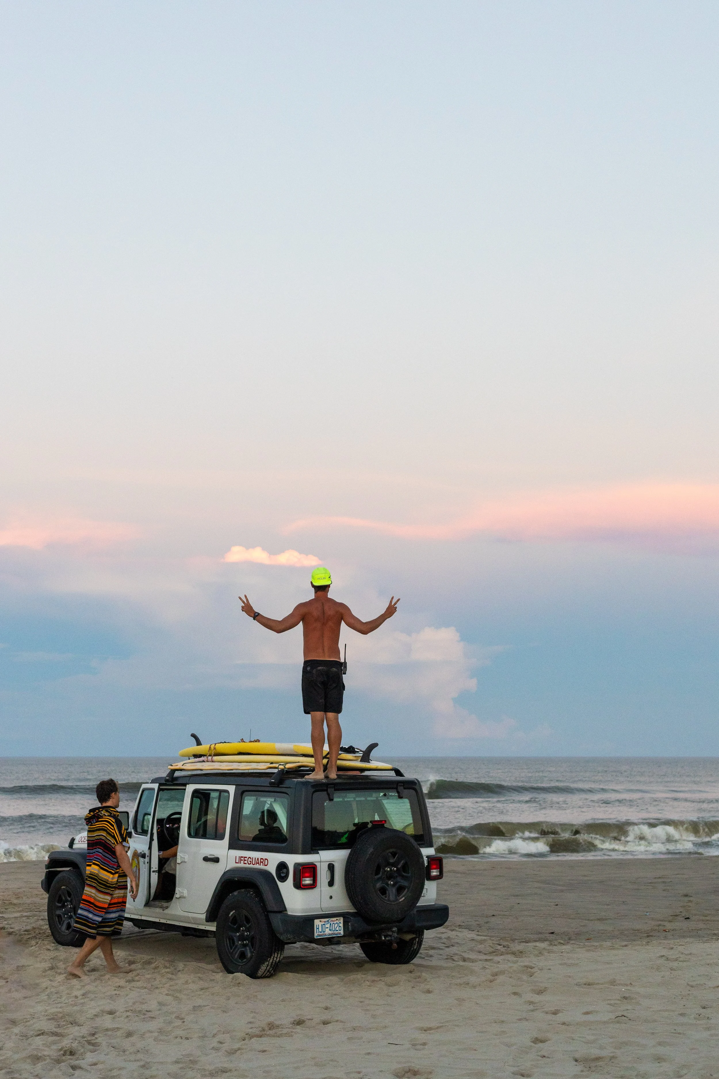 A shirtless man wearing a yellow swim cap and black shorts stands on top of a parked white police SUV on a sandy beach, making peace signs with both hands. A woman wrapped in a colorful beach towel walks towards the vehicle, and the ocean waves are v